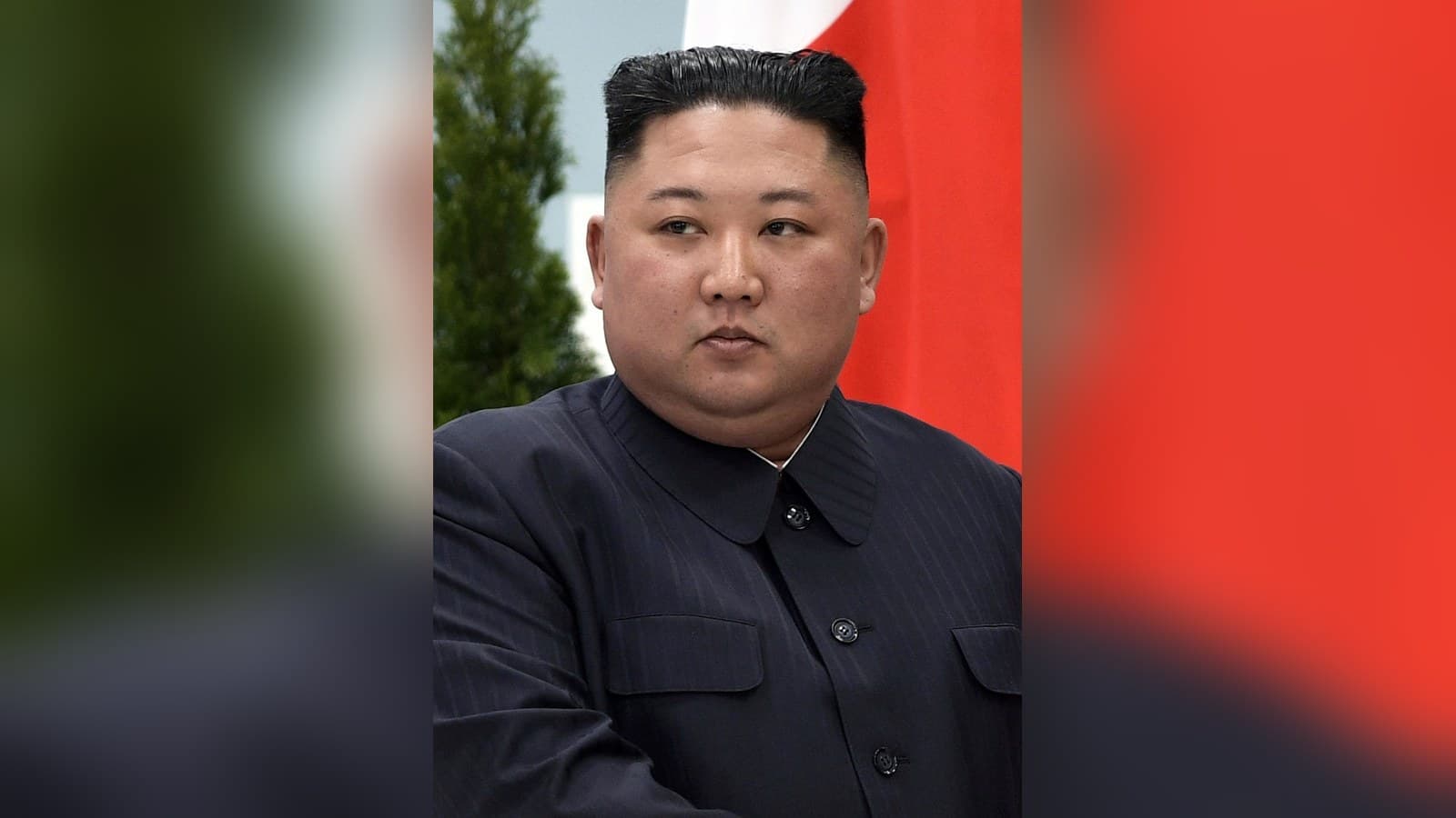 North Korean leader Kim Jong Un in a dark Mao-style suit, standing in front of a red flag and a fir tree in Vladivostok in 2019