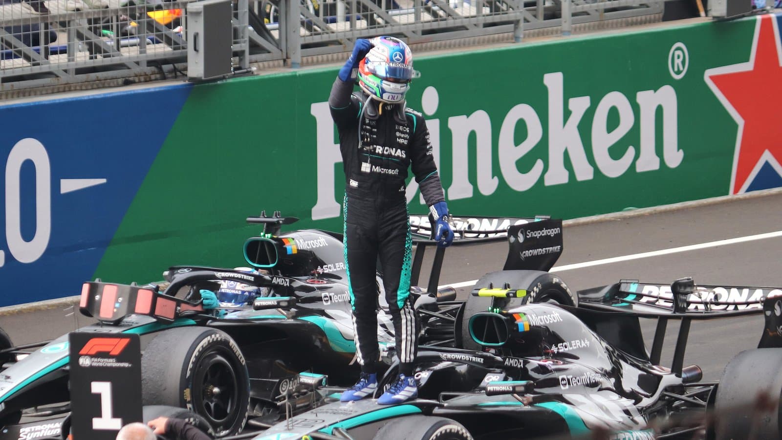 Mercedes driver Kimi Antonelli standing on his silver and black Formula 1 car with one arm raised, in front of a green Heineken advertising board.