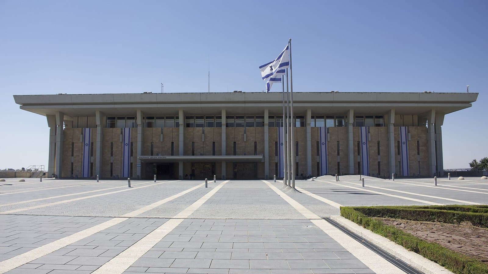 The front of the Knesset building in Jerusalem, a low rectangular stone building with tall columns and an Israeli flag flying above a wide paved plaza.