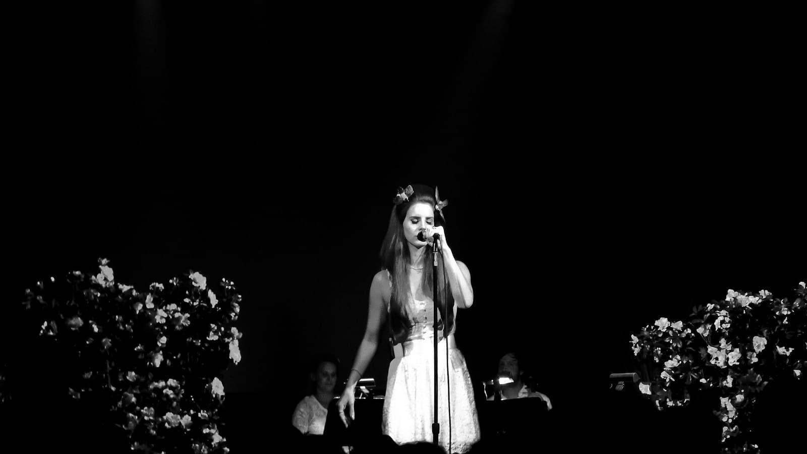 Black-and-white photo of Lana Del Rey in a white dress singing at a microphone stand on a dark stage framed by flowers.