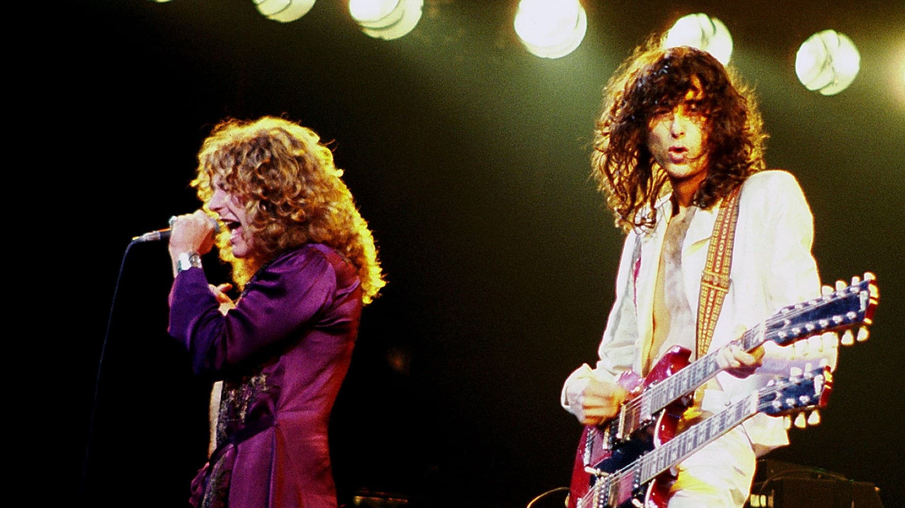 Robert Plant in a purple shirt singing into a microphone beside Jimmy Page playing a red double-neck guitar under bright stage lights, 1977.