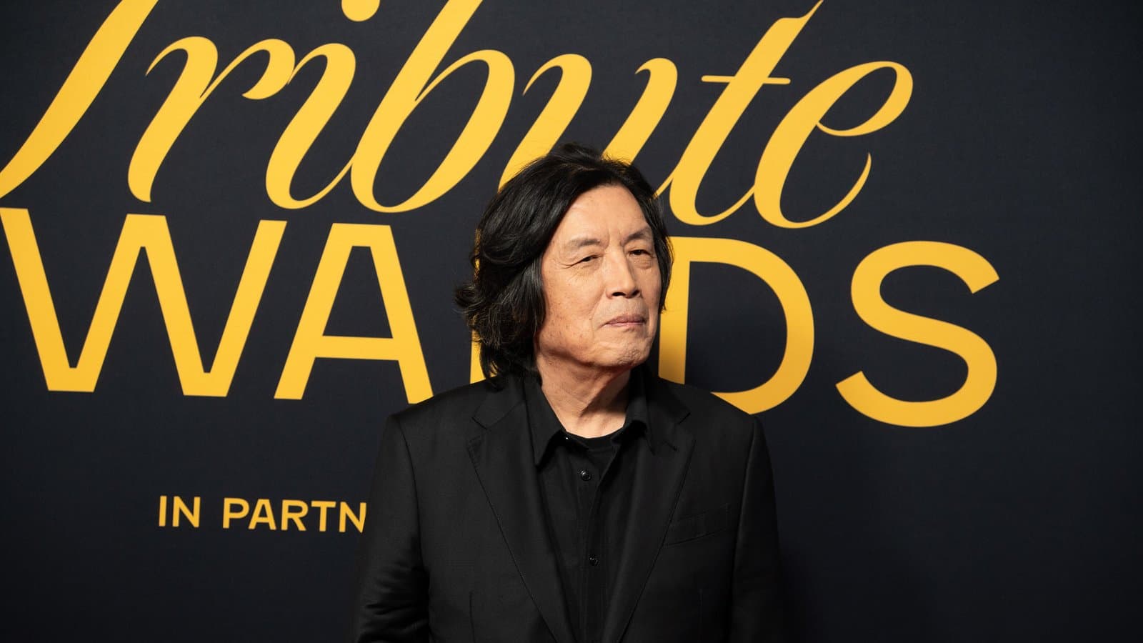 Director Lee Chang-dong, with shoulder-length dark hair and a black jacket, stands in front of a black-and-gold Tribute Awards backdrop.