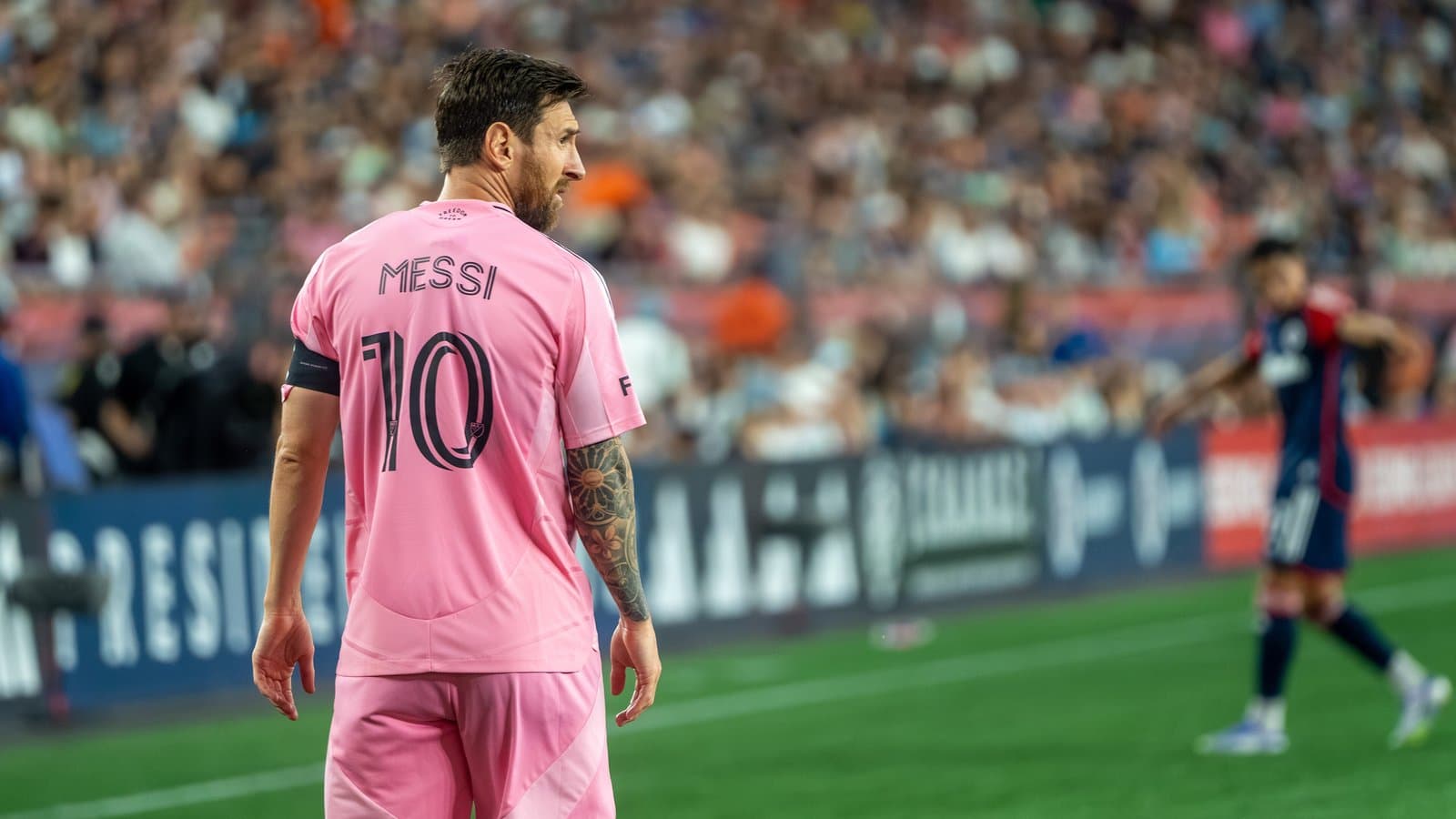 Lionel Messi in a pink Inter Miami No. 10 jersey and black shorts on the field during a match, with fans in the stands behind him.