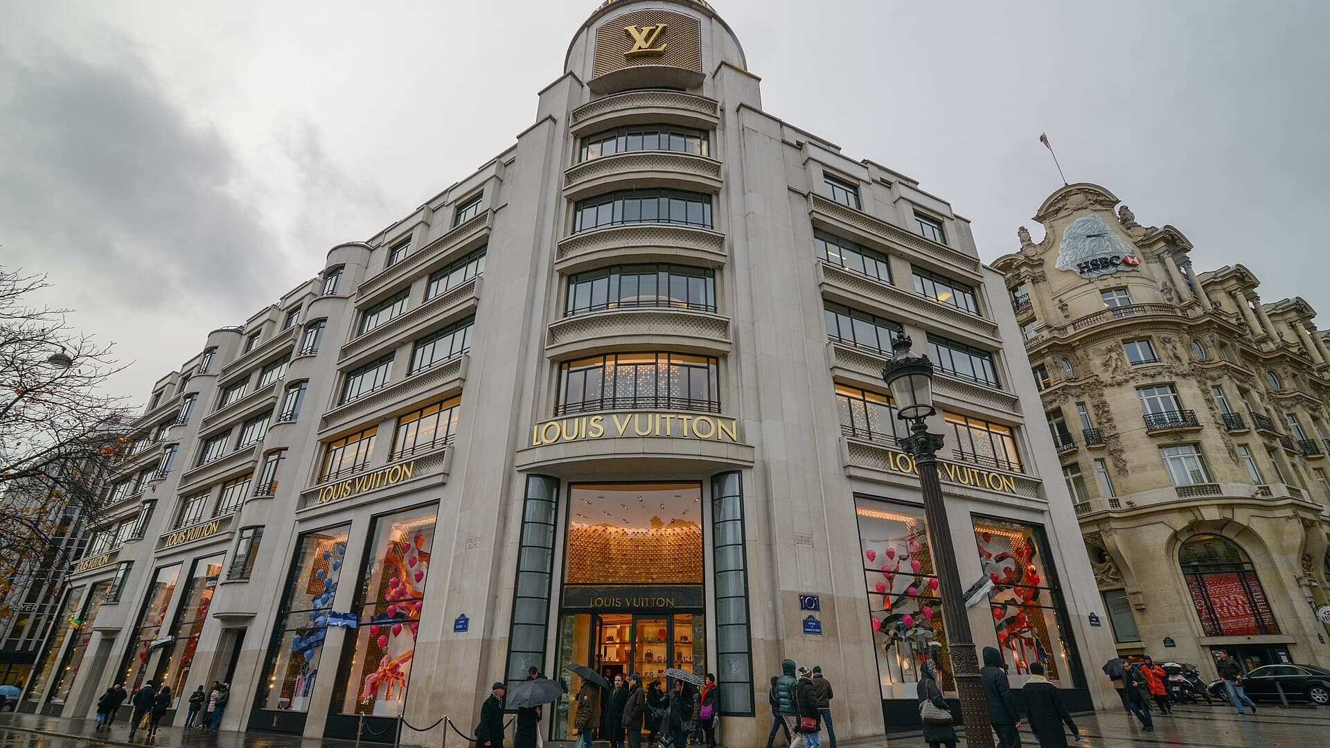 The cream stone Louis Vuitton flagship building on a Paris street corner under a grey sky, gold Louis Vuitton lettering above lit shop windows and shoppers with umbrellas on the pavement.