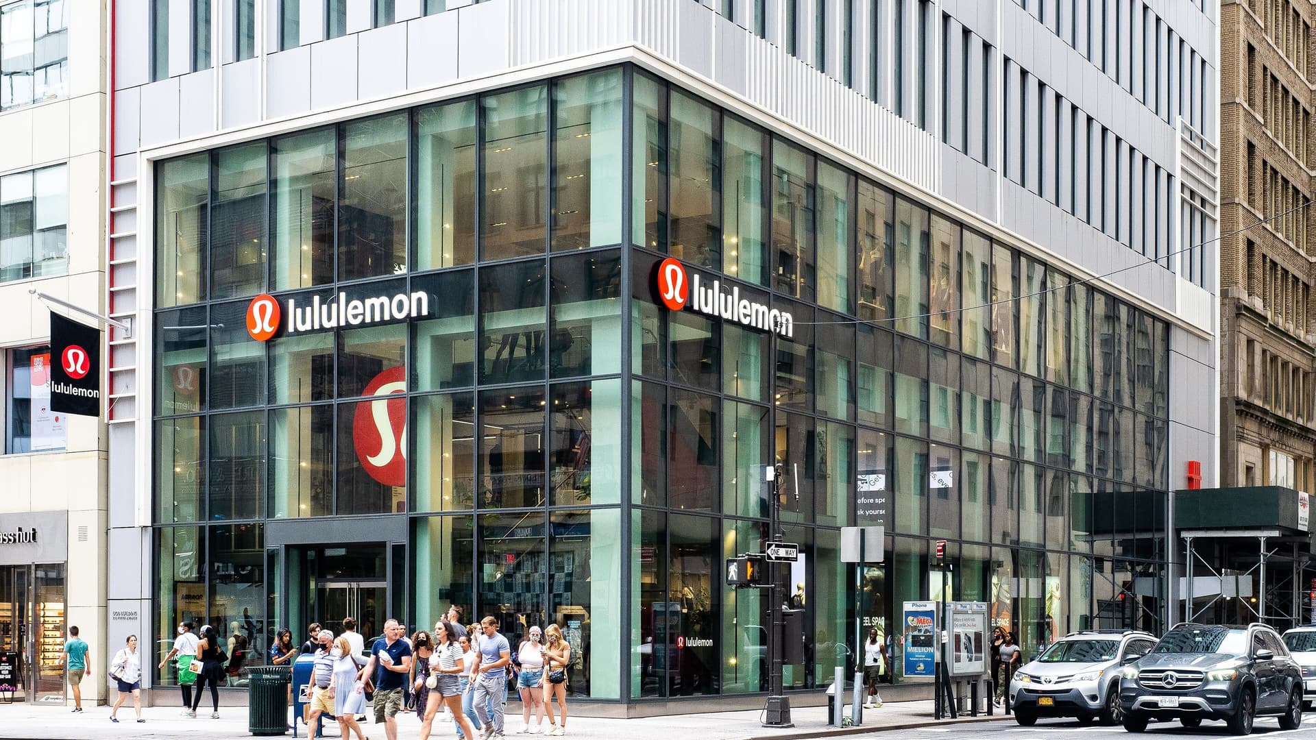 Corner lululemon store with tall glass windows and red lululemon logos, with shoppers walking along the sidewalk outside.