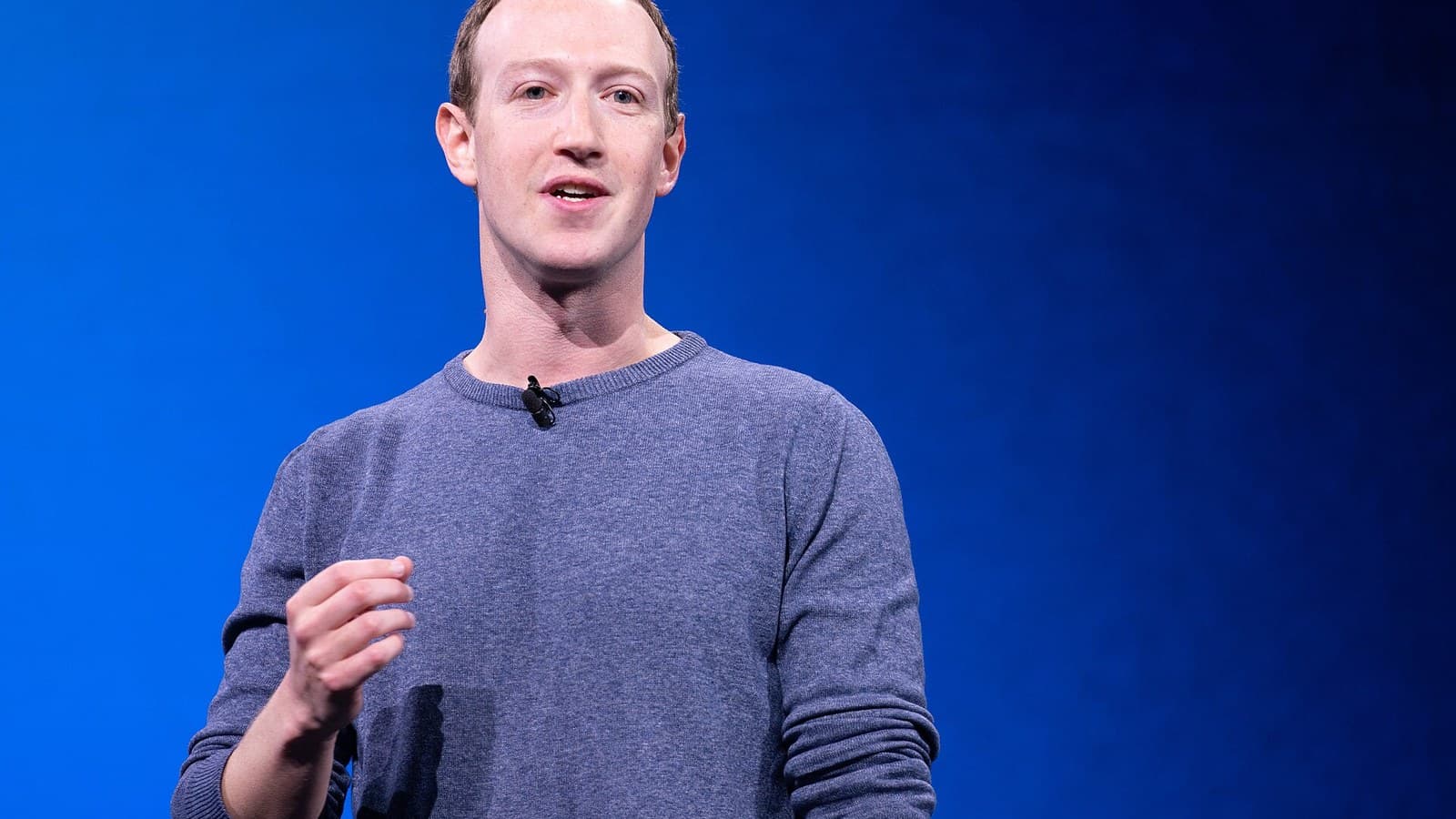 Mark Zuckerberg in a blue crewneck sweater speaking on stage against a bright blue background