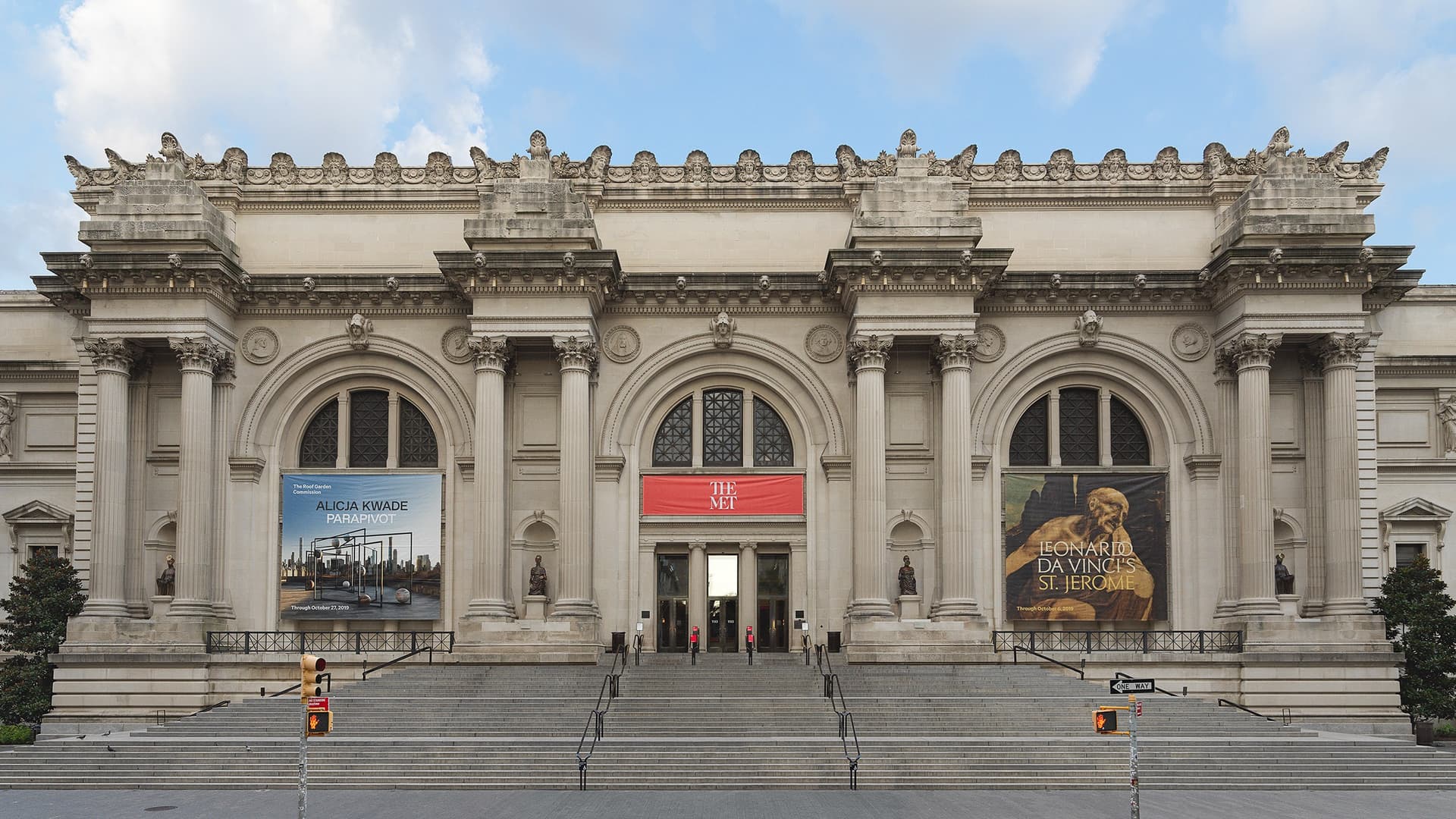 The Beaux-Arts facade of the Metropolitan Museum of Art on Fifth Avenue, with columns, three arched windows, a red 'The Met' banner over the entrance and wide stone steps.