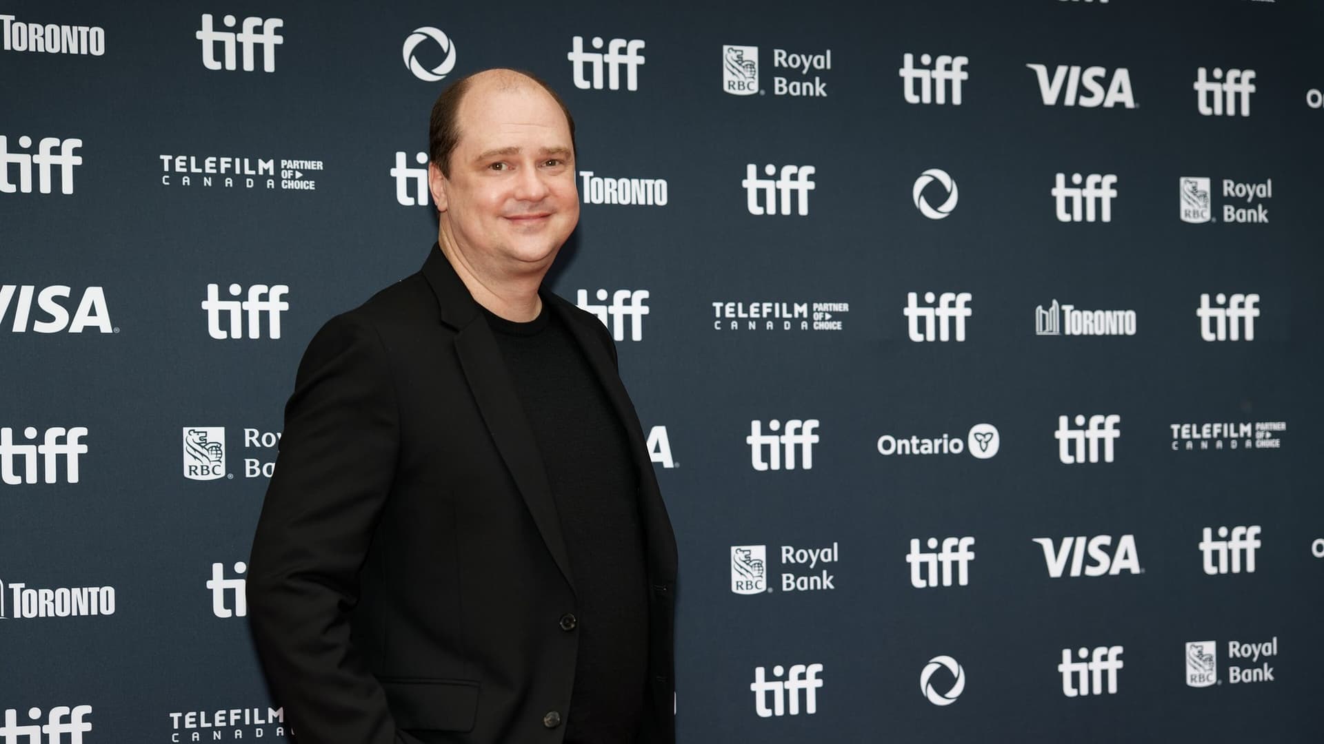 Director Mike Flanagan, wearing a black suit and black shirt, smiling on a red carpet in front of a TIFF sponsor backdrop.