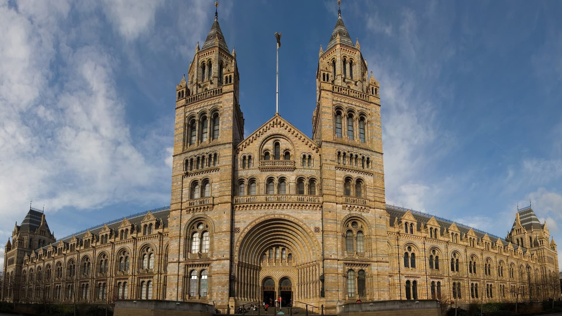 The Romanesque terracotta facade of the Natural History Museum in London, with its two central towers and arched entrance, under a blue sky with clouds.