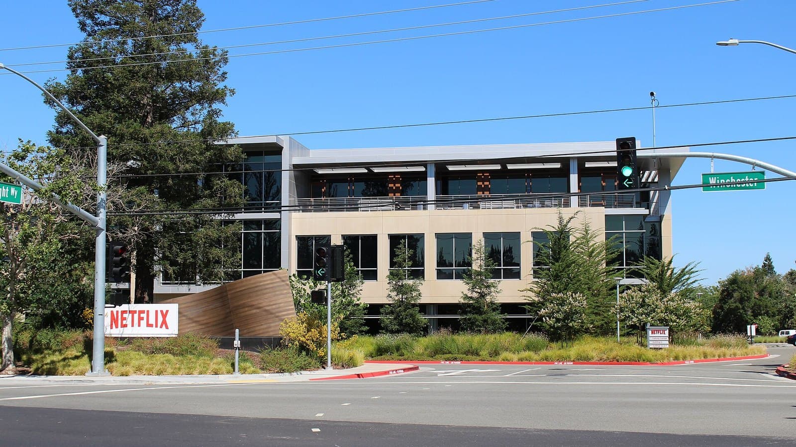 A modern three-storey office building with large windows behind trees and landscaping on a street corner, with a red Netflix sign on a low wall in the foreground under a clear sky.