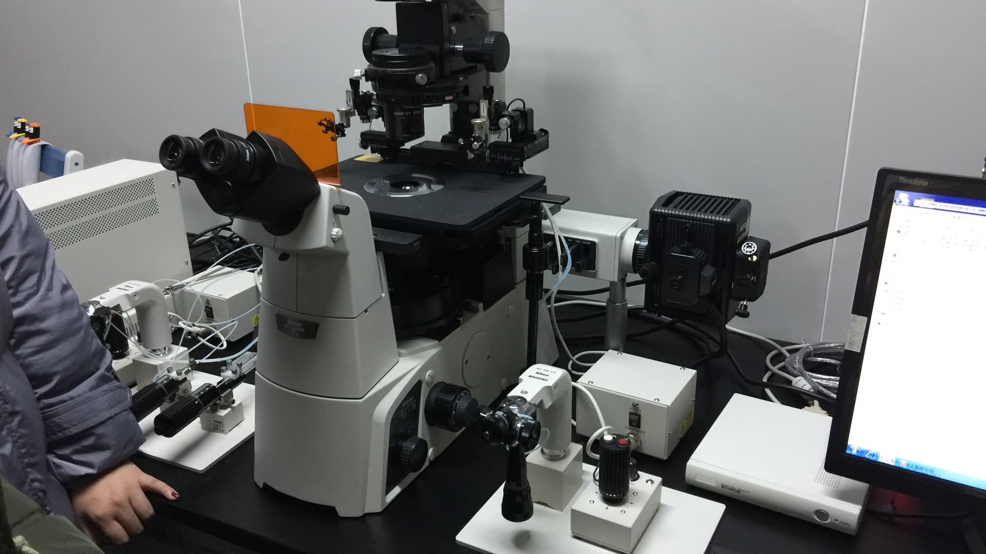 A white Nikon inverted research microscope with eyepieces, a camera module and cabling on a lab bench next to a computer monitor.