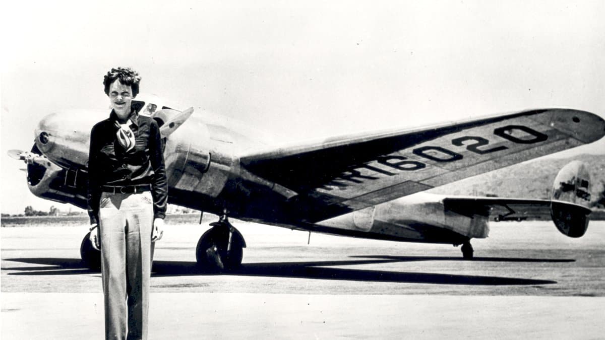 Black-and-white photo of Amelia Earhart standing in front of her silver twin-engine Lockheed Electra, registration NR16020 painted on the wing.