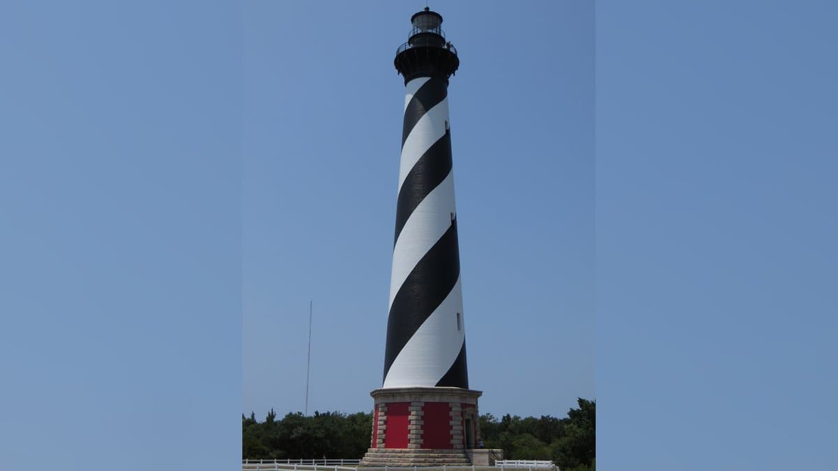 The black-and-white spiral-striped Cape Hatteras Lighthouse rising above green shrubs under a clear blue sky.