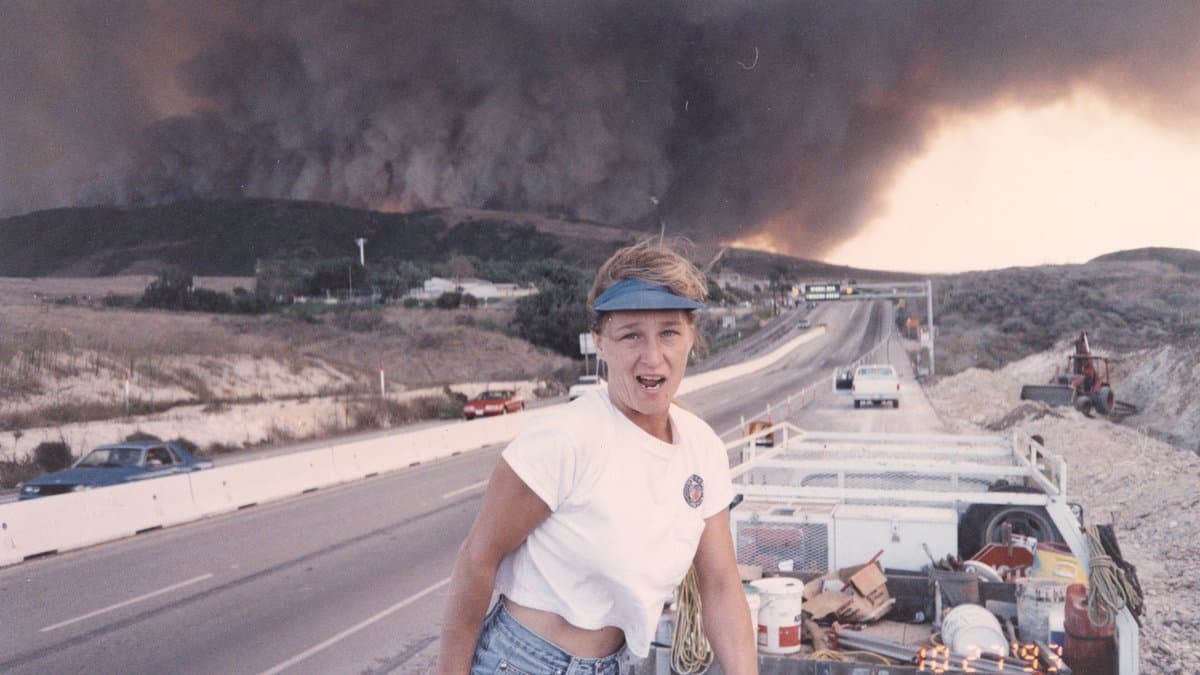 A woman in a white T-shirt and blue visor stands beside a truck on a highway as a massive wall of dark wildfire smoke rises over the hills behind her.