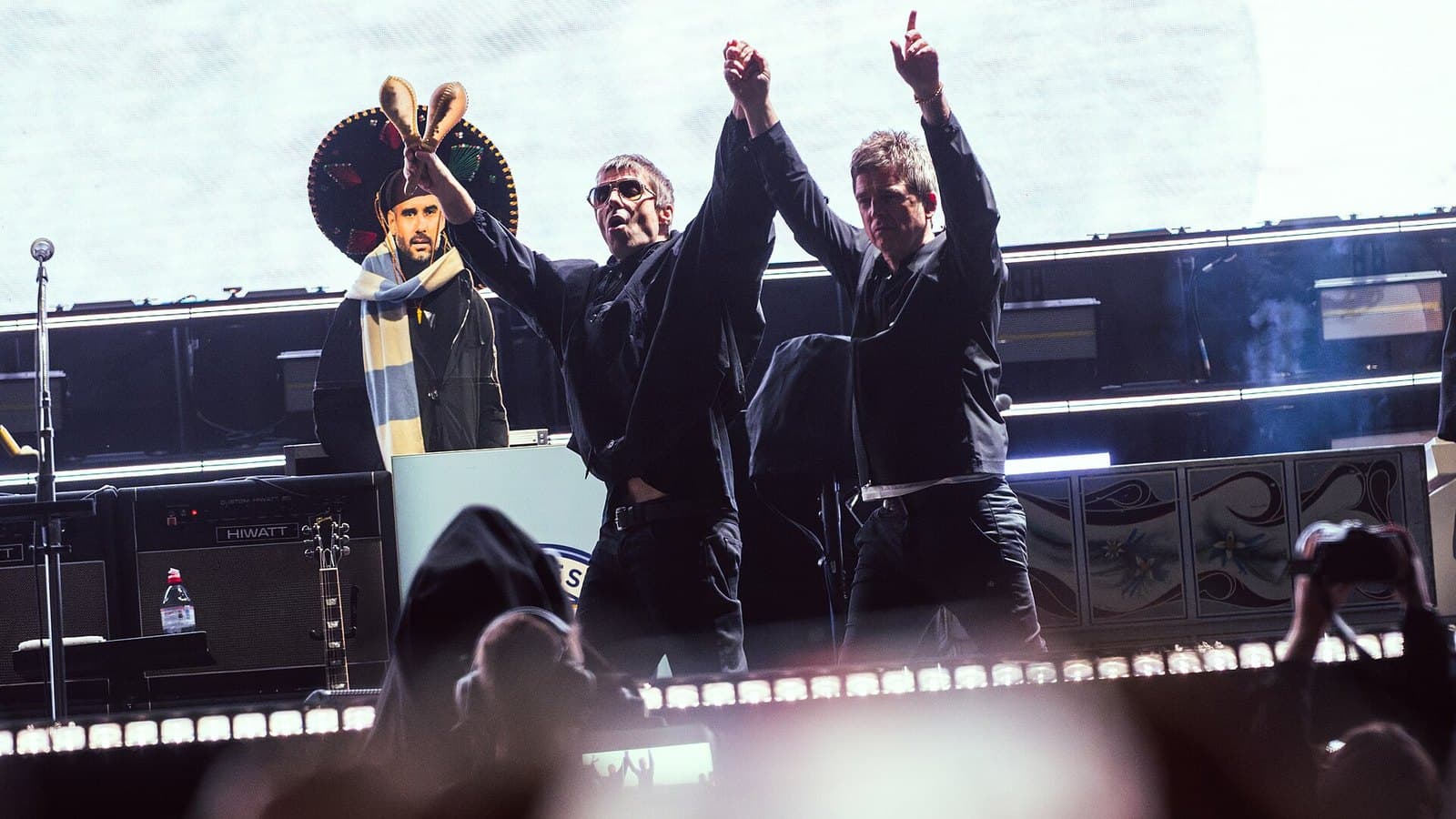 Liam and Noel Gallagher, dressed in black, raise their joined hands on a stadium stage as phone lights glow from the crowd in front of them.