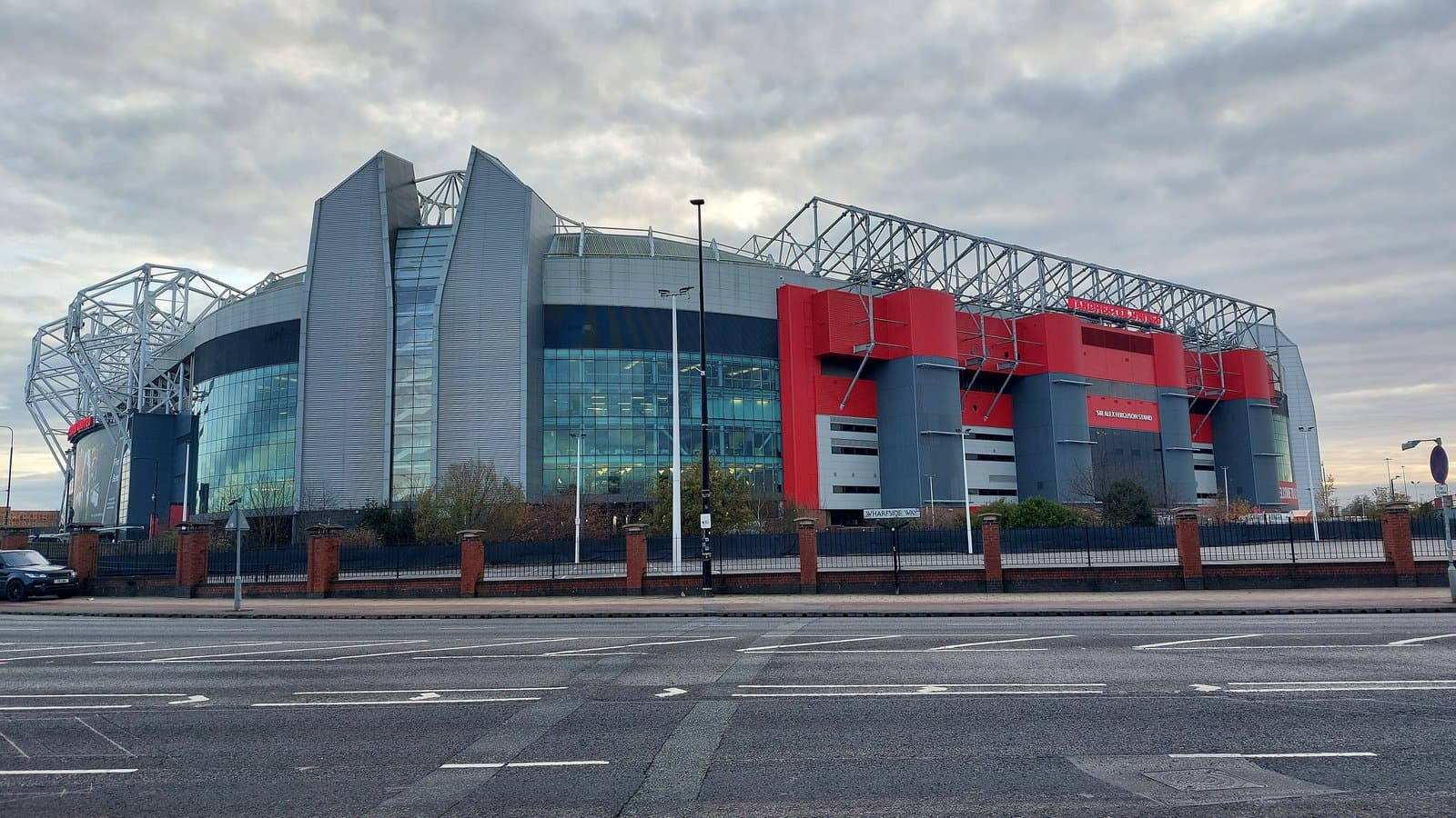 Exterior of Old Trafford stadium in Manchester with red cladding and a glass-fronted stand under a pale sky.
