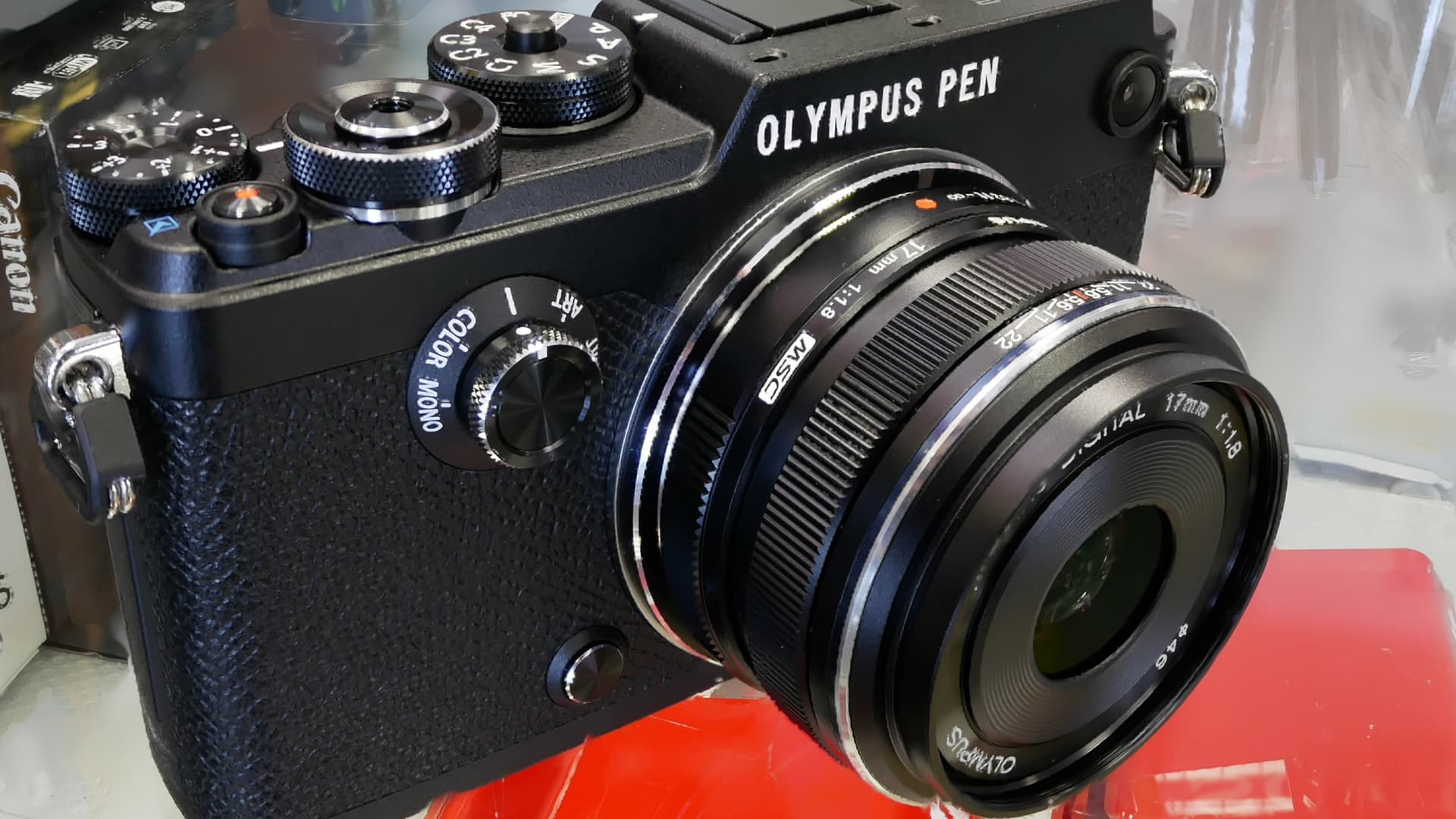 A black Olympus PEN-F mirrorless camera with knurled metal dials, a color and monochrome selector on the front, and a 17mm f/1.8 lens.