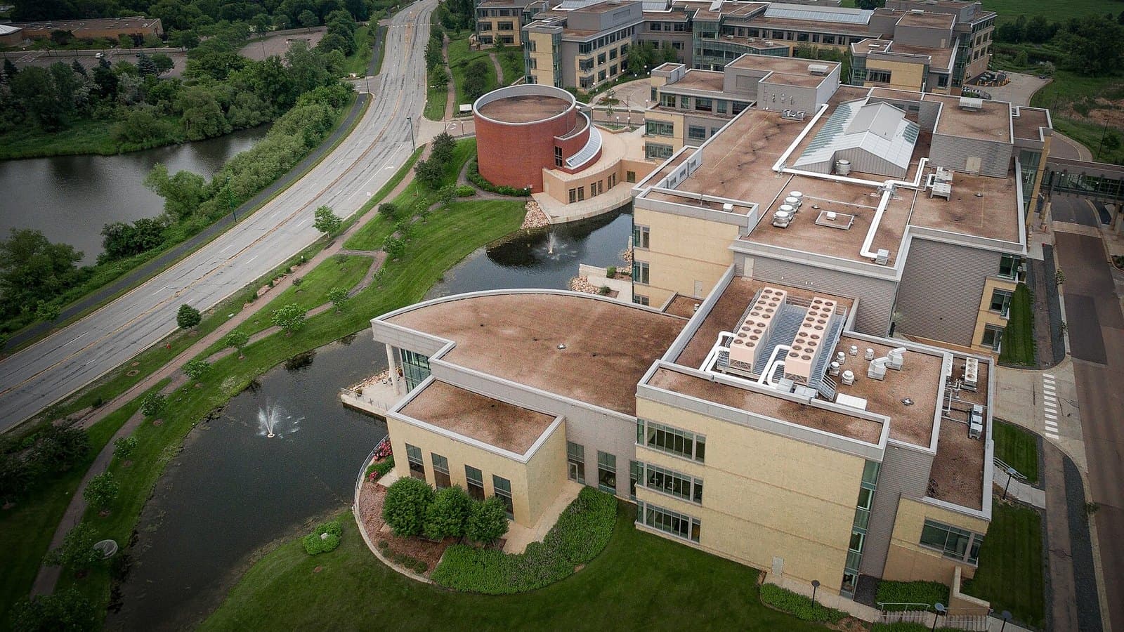 Aerial view of a large beige office campus with flat roofs, a round building, a pond and a curving road lined with green trees and lawns.