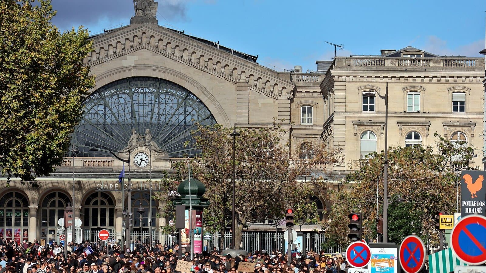 A large crowd of demonstrators filling the street in front of the Gare du Nord railway station in Paris, with its glass arches and stone statues above.