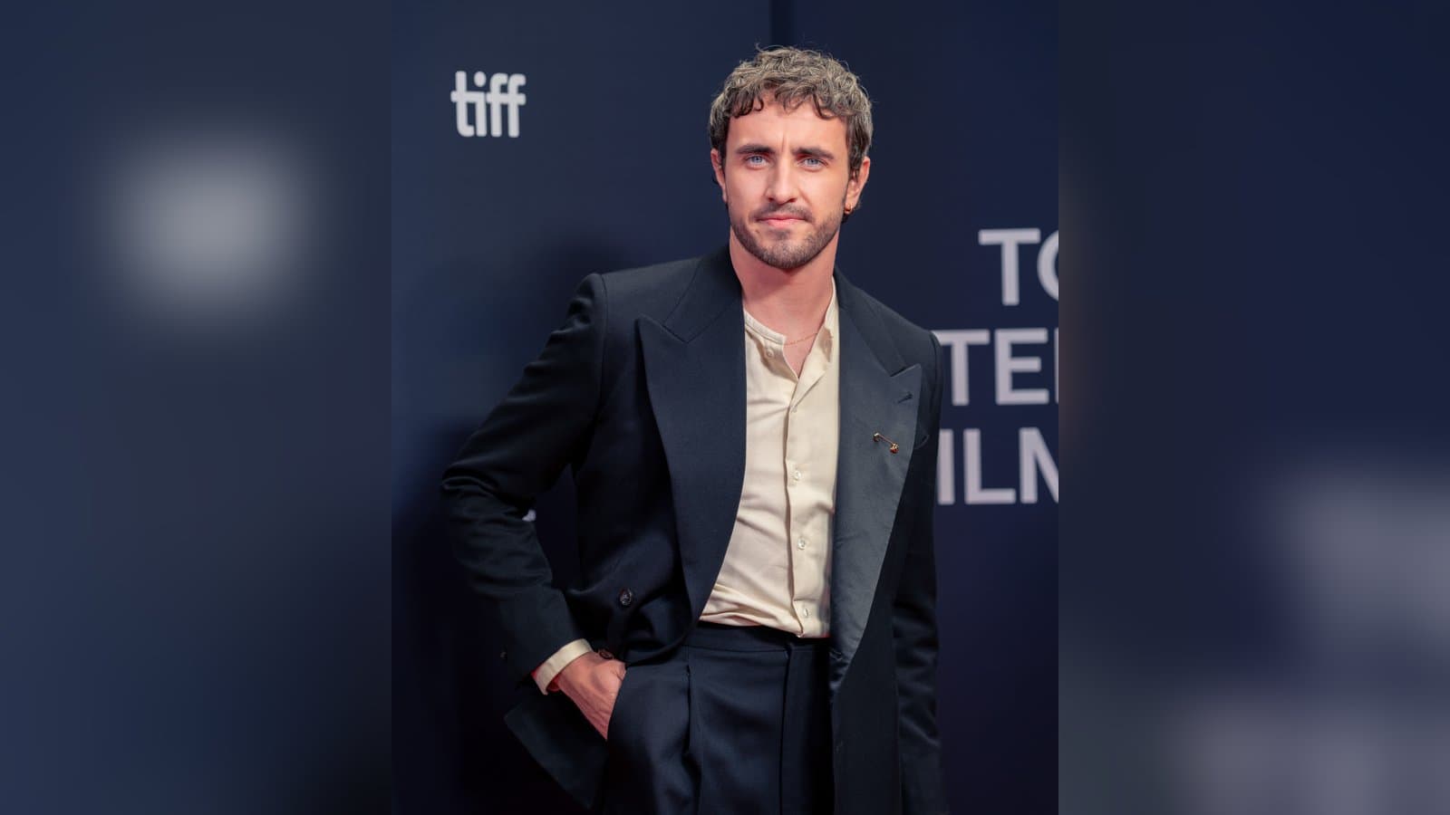 Paul Mescal, with curly hair and a beard, in a black suit and cream shirt, poses in front of a TIFF backdrop.