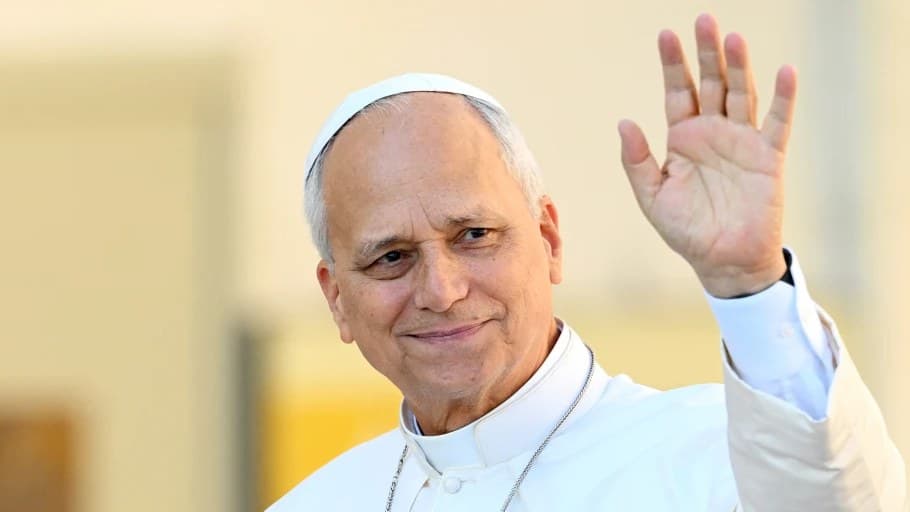 Pope Leo XIV in white cassock and zucchetto, smiling and waving with his left hand.