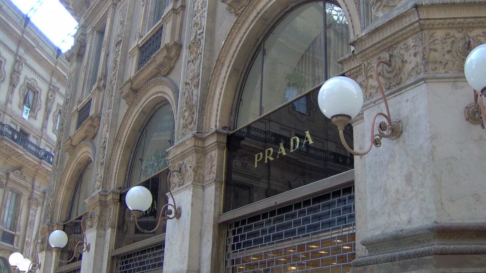Ornate stone arches of the Galleria Vittorio Emanuele II in Milan, with gold PRADA lettering on black glass above the boutique's shuttered windows and globe lamps on curved brackets.