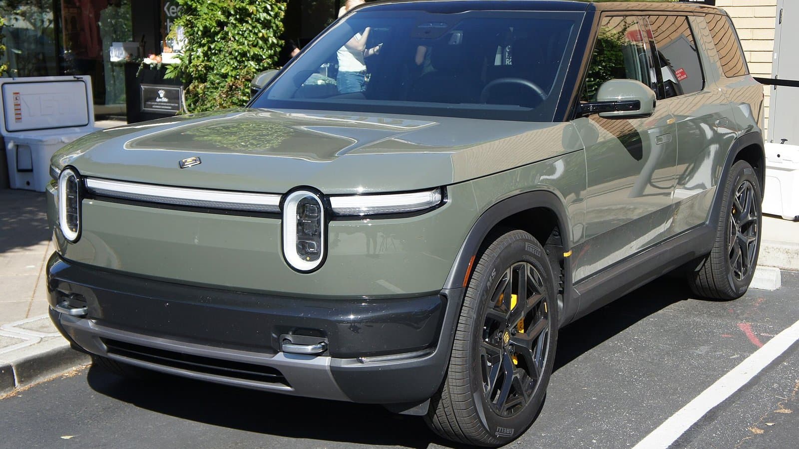 A sage green Rivian R2 electric SUV with oval headlights and a light bar, parked on a street.