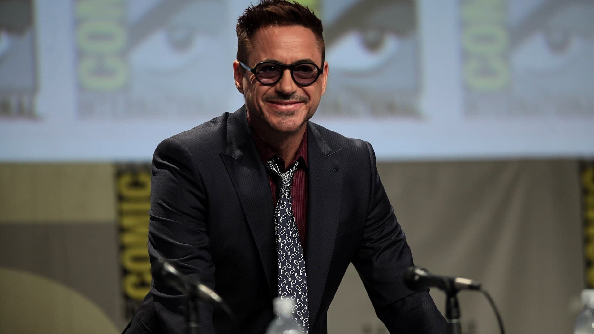 Robert Downey Jr. in round dark glasses and a dark suit smiling at a microphone on a Comic-Con panel stage.