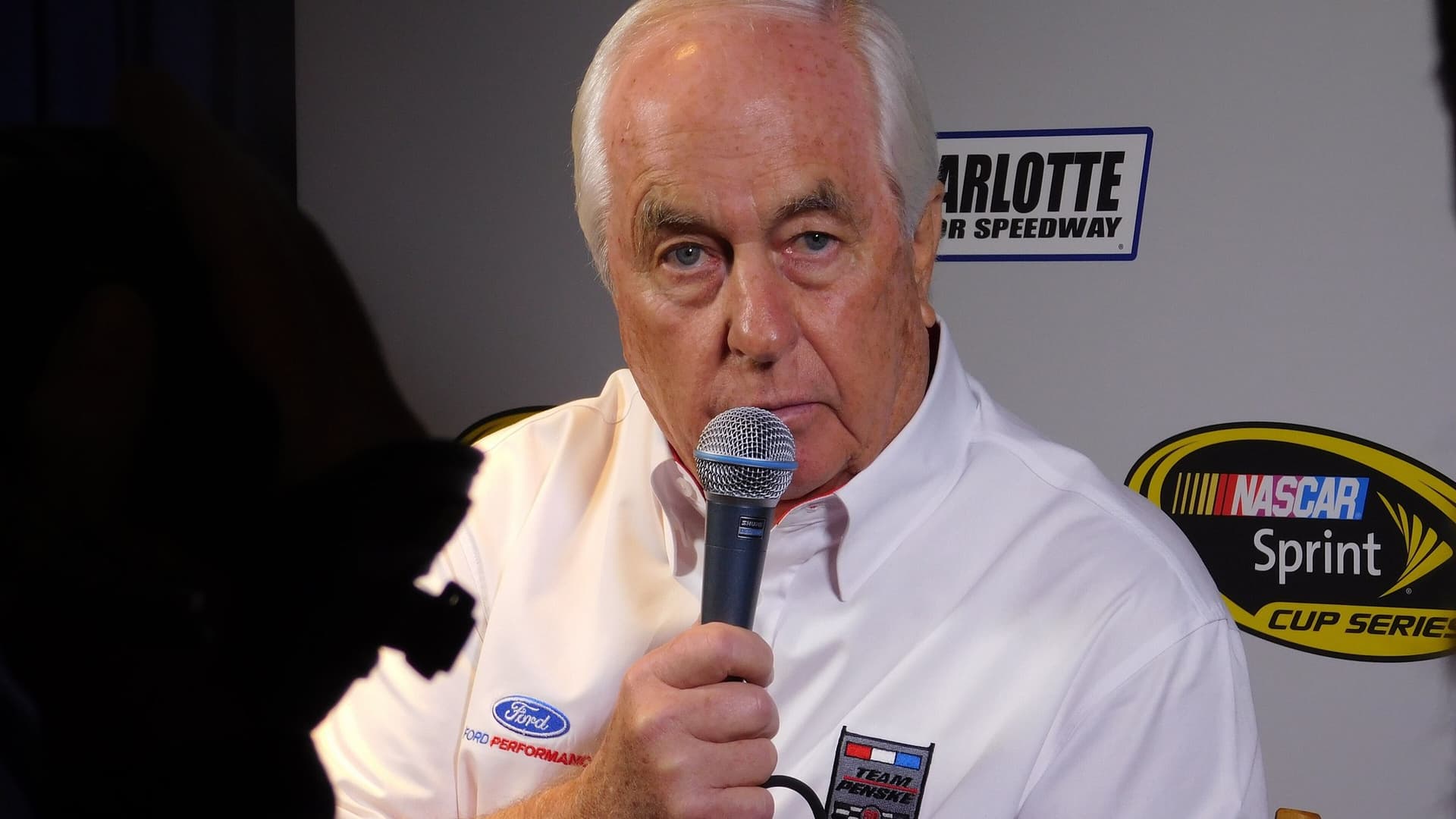 Roger Penske, white-haired and wearing a white Team Penske shirt, speaks into a handheld microphone in front of a NASCAR backdrop.