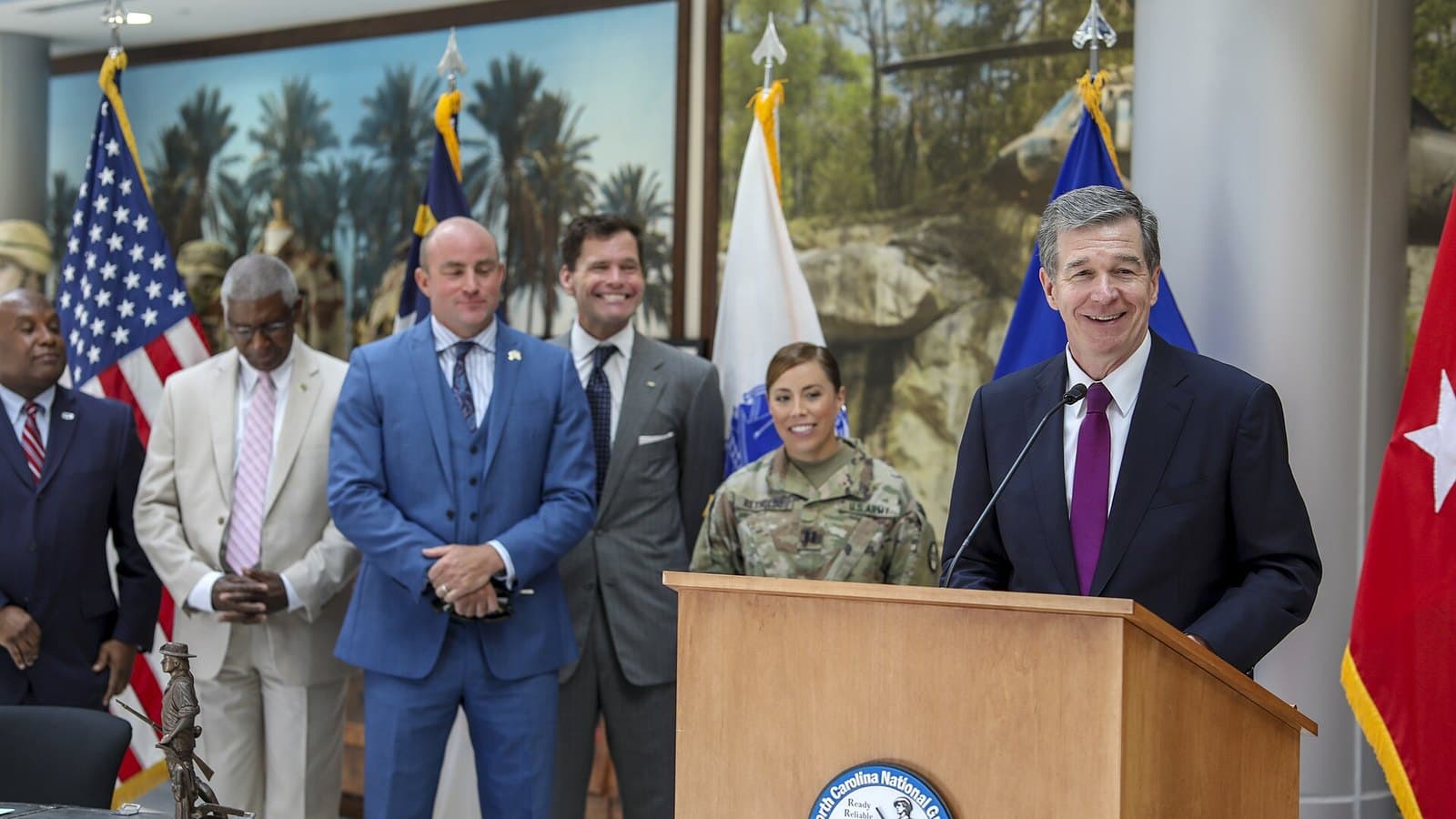 Roy Cooper, in a dark suit, speaks into a microphone at an outdoor bill-signing ceremony with uniformed North Carolina National Guard members behind him.