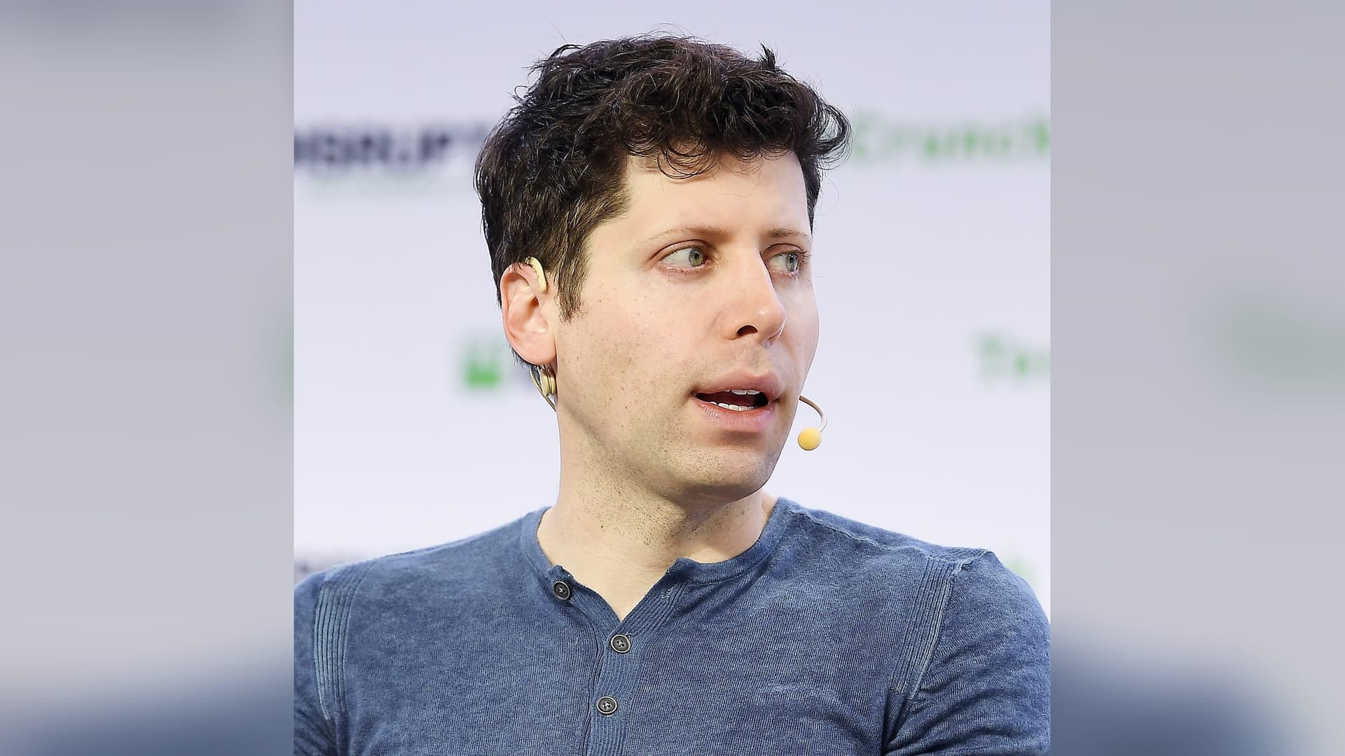 OpenAI CEO Sam Altman, wearing a blue henley and a headset microphone, speaking on stage at TechCrunch Disrupt in San Francisco.