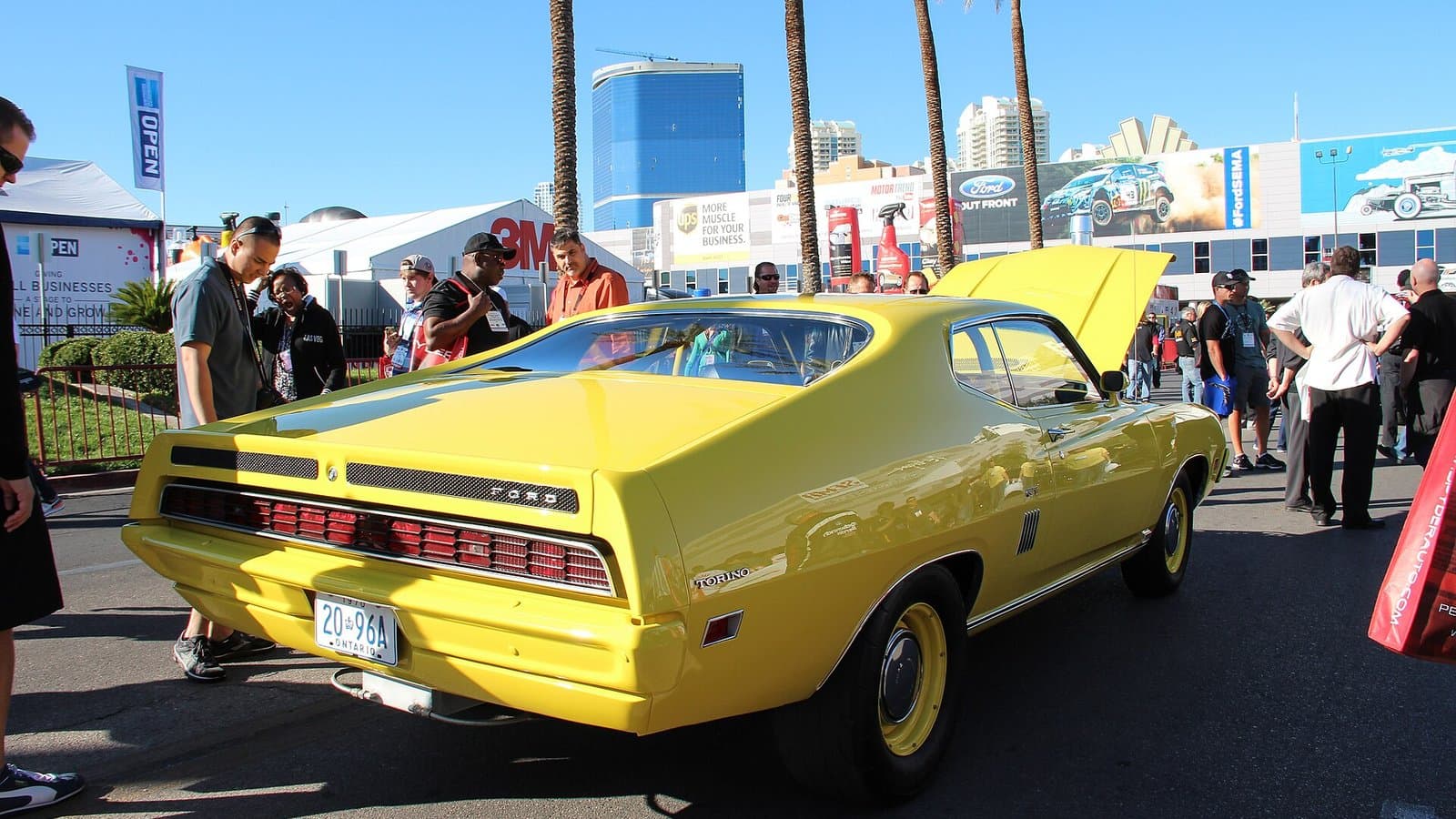 Crowds walking past a yellow classic muscle car with its hood up at an outdoor display at the SEMA Show in Las Vegas.