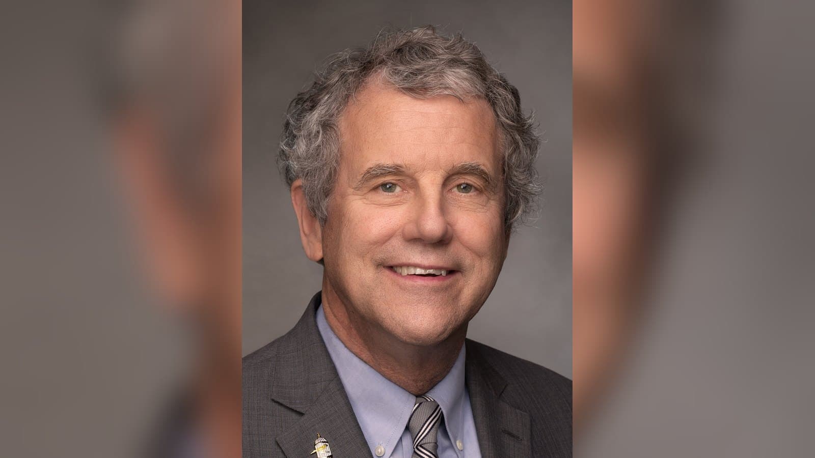 Official portrait of Sherrod Brown, with curly gray hair, in a gray suit and striped tie, smiling against a dark backdrop.