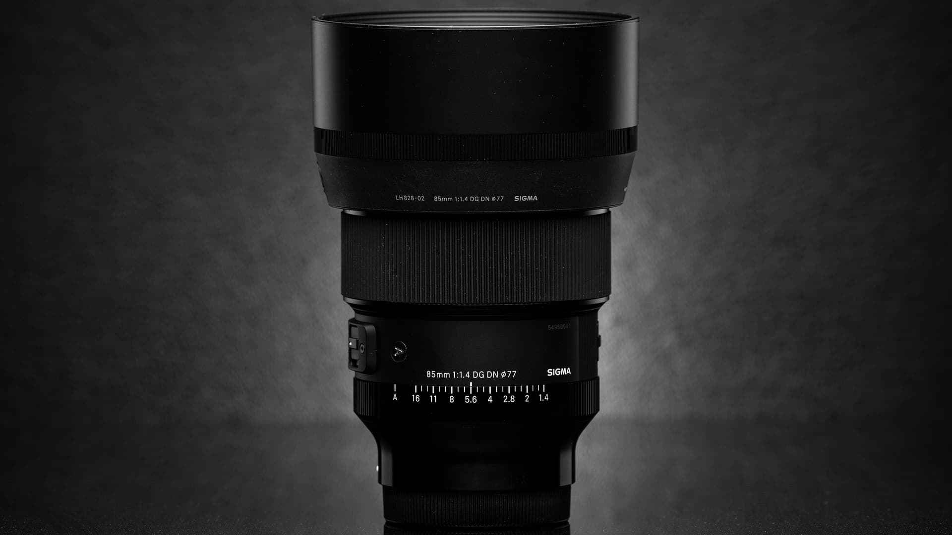 A black Sigma 85mm F1.4 DG DN Art lens with its hood attached, standing upright against a dark grey background, with the aperture ring markings visible.
