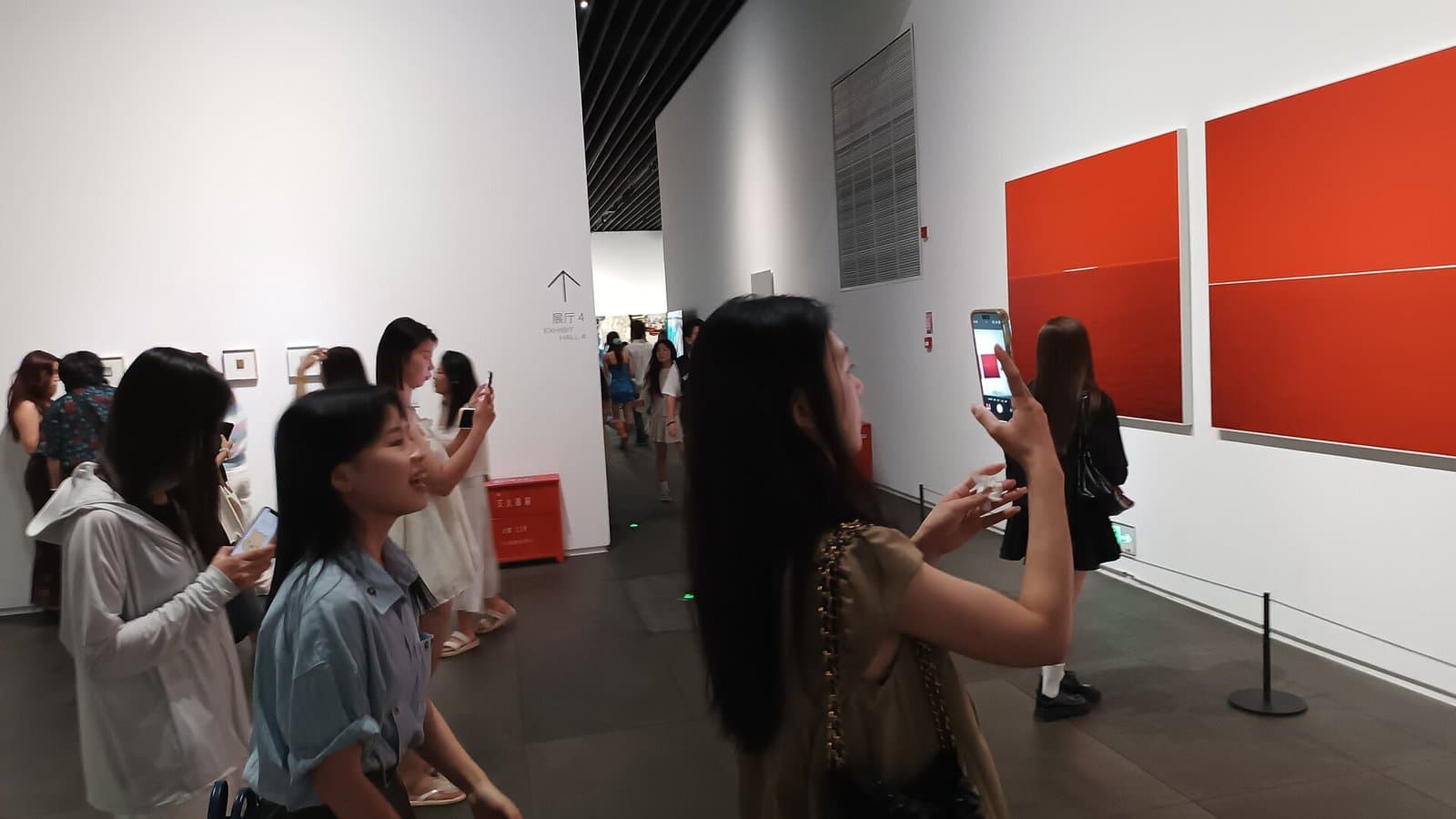 Several women holding up smartphones to photograph large red paintings on the white walls of an art museum gallery.