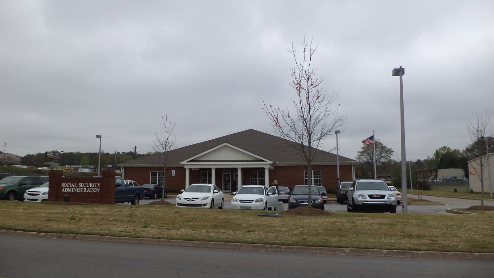 A single-storey brick Social Security Administration office with a white-columned entrance, cars in the parking lot and a sign reading Social Security Administration on the lawn under a grey sky.
