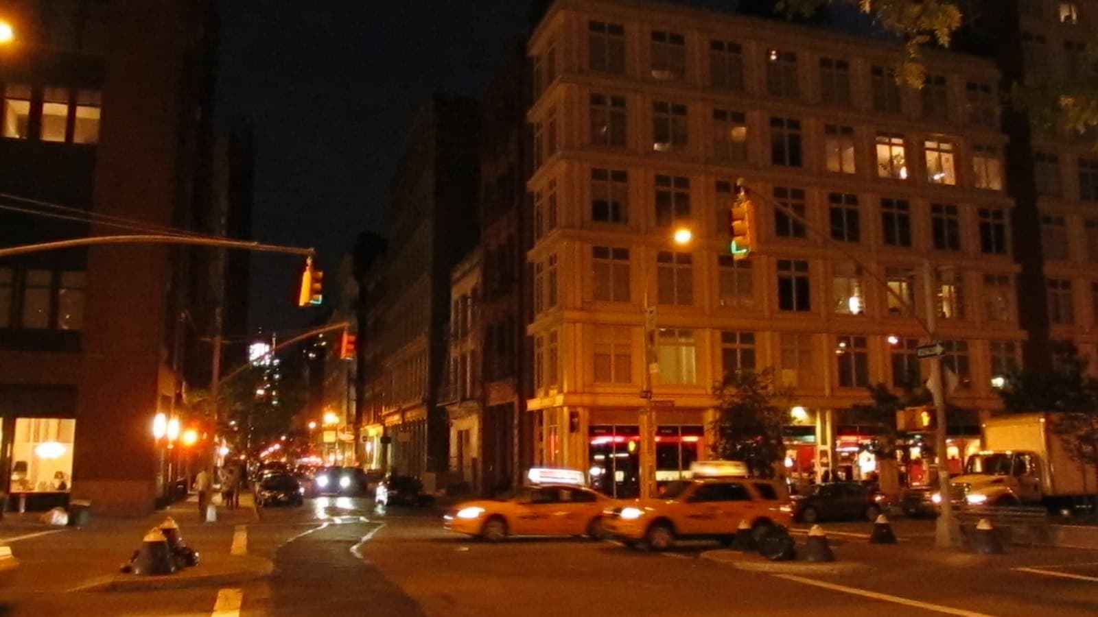 A SoHo street intersection in New York at night, with yellow taxis, streetlights and lit storefronts beneath tall brick and cast-iron buildings.