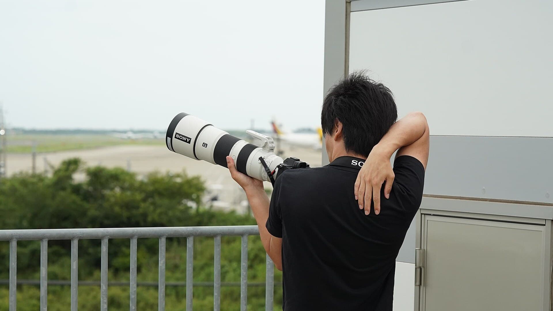 A man in a black Sony shirt holds a camera with a long white Sony telephoto lens up to his eye on an observation deck overlooking an airport runway.