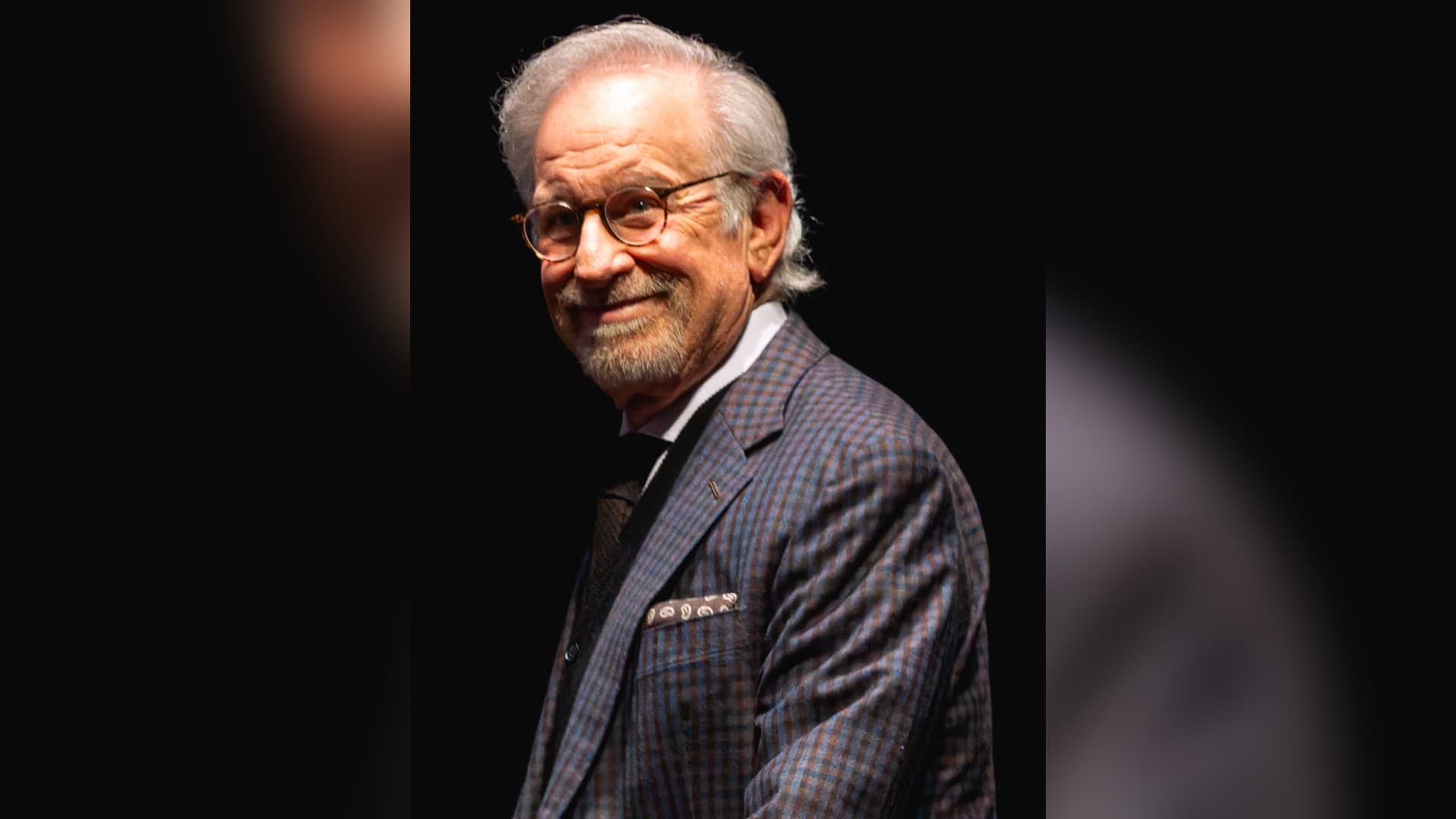 Steven Spielberg, grey-haired with round tortoiseshell glasses and a checked blazer, smiling over his shoulder against a black background.