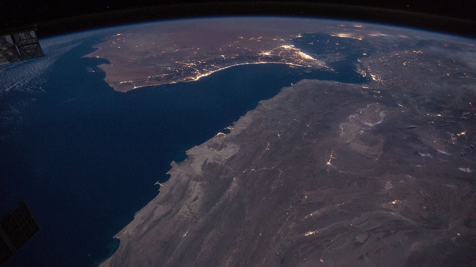 A night view from space of the Strait of Hormuz, with the dark mass of the Musandam peninsula and the lights of Gulf cities along the coast.
