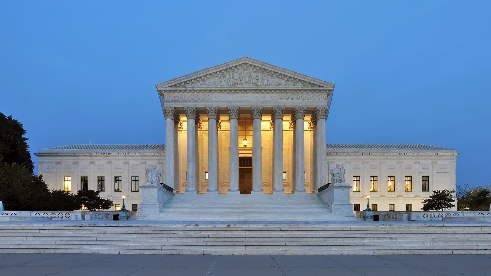 The white marble U.S. Supreme Court Building in Washington, D.C., lit from within at dusk, with its columned front and wide plaza under a blue sky.