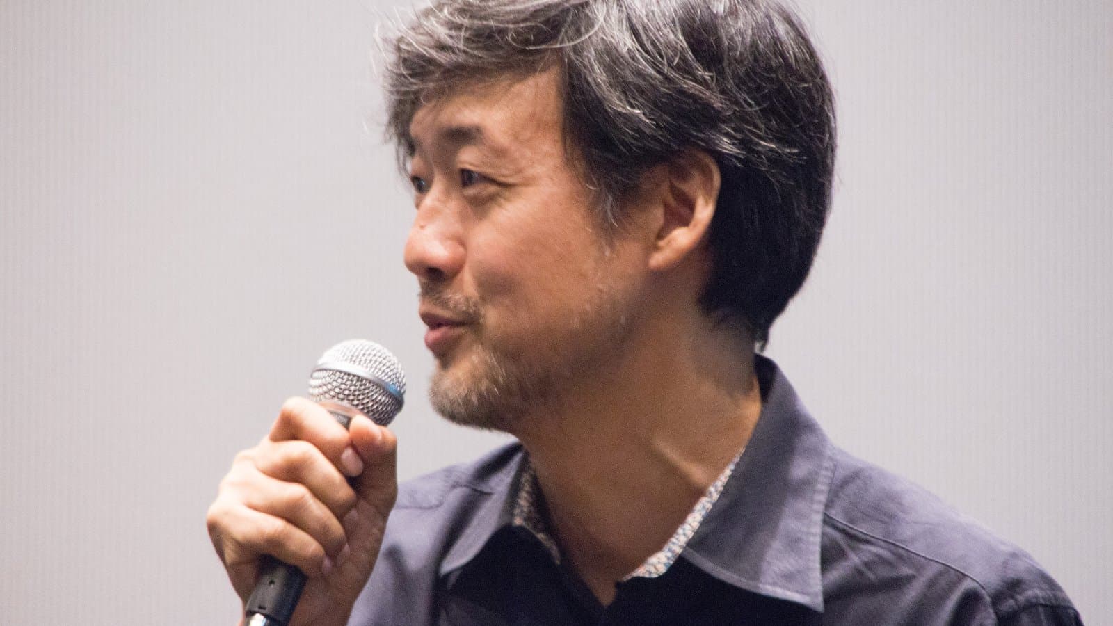 Japanese director Takashi Yamazaki, with grey-streaked hair and a short beard, speaks into a microphone in front of a plain backdrop.