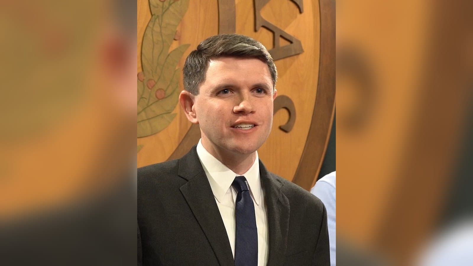 James Talarico, in a dark suit and tie, smiles at a press conference inside the Texas Capitol in Austin, with an ornate seal behind him.