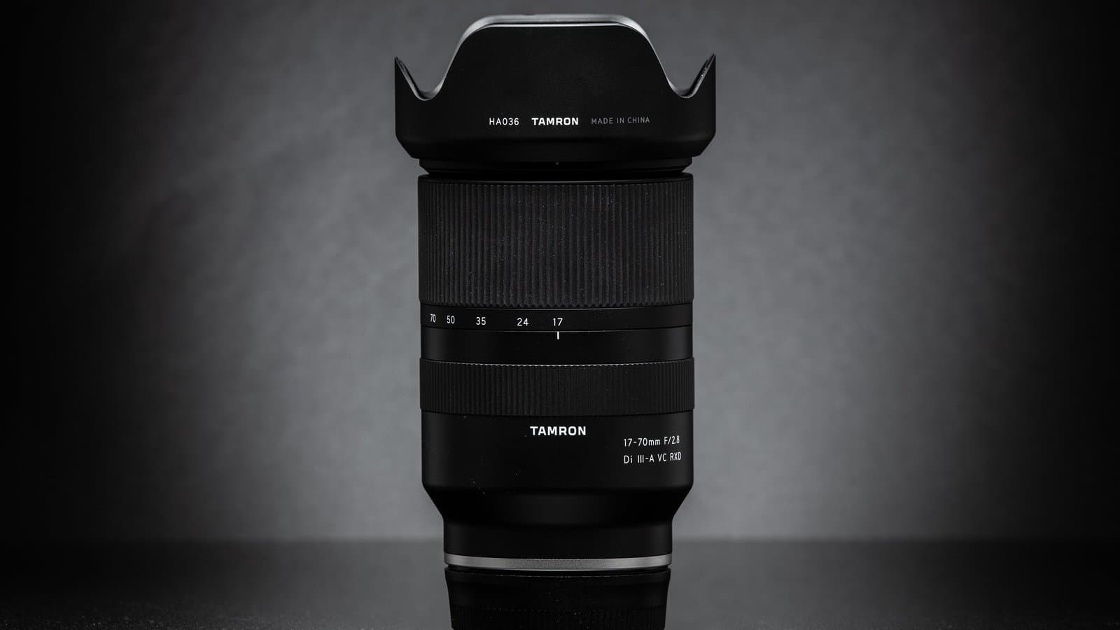 A black Tamron 17-70mm f/2.8 zoom lens with its petal-shaped hood attached, standing upright against a dark gray background.