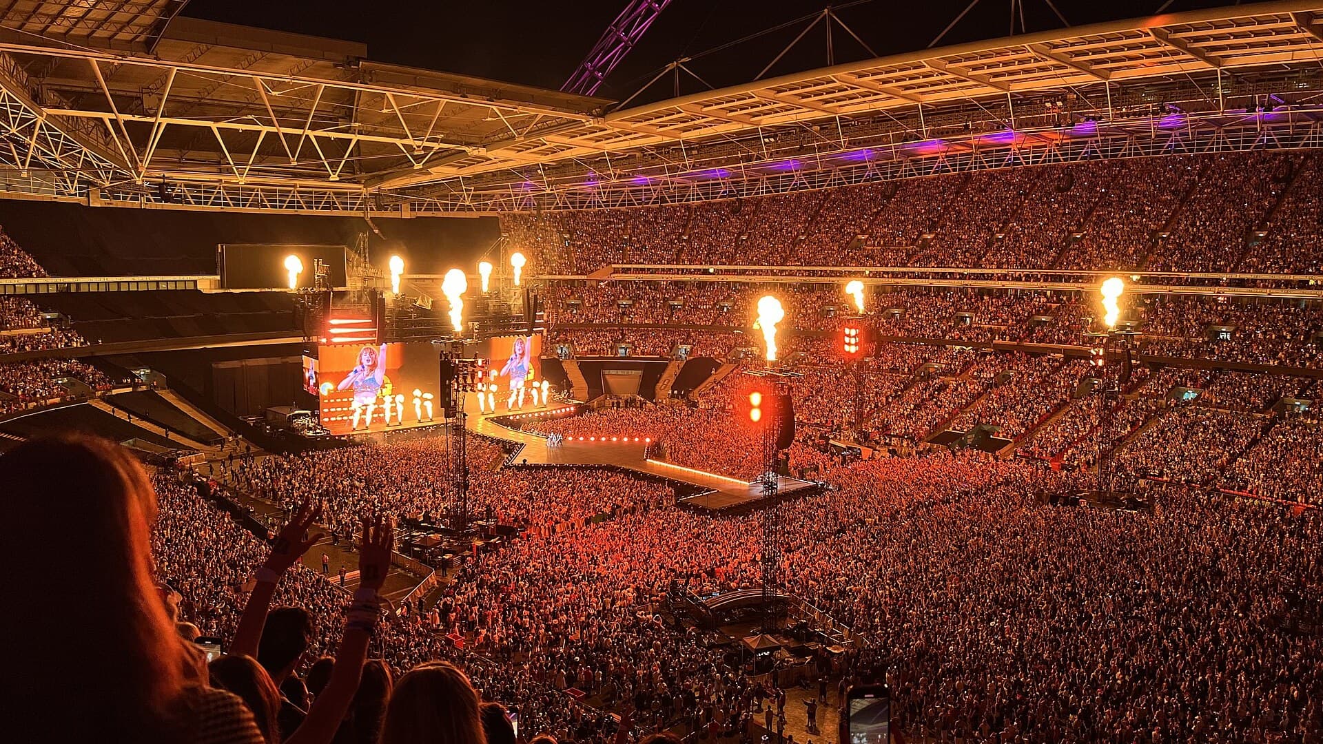 A packed Wembley Stadium at night lit orange by stage fireworks, with Taylor Swift shown on giant video screens during an Eras Tour concert.
