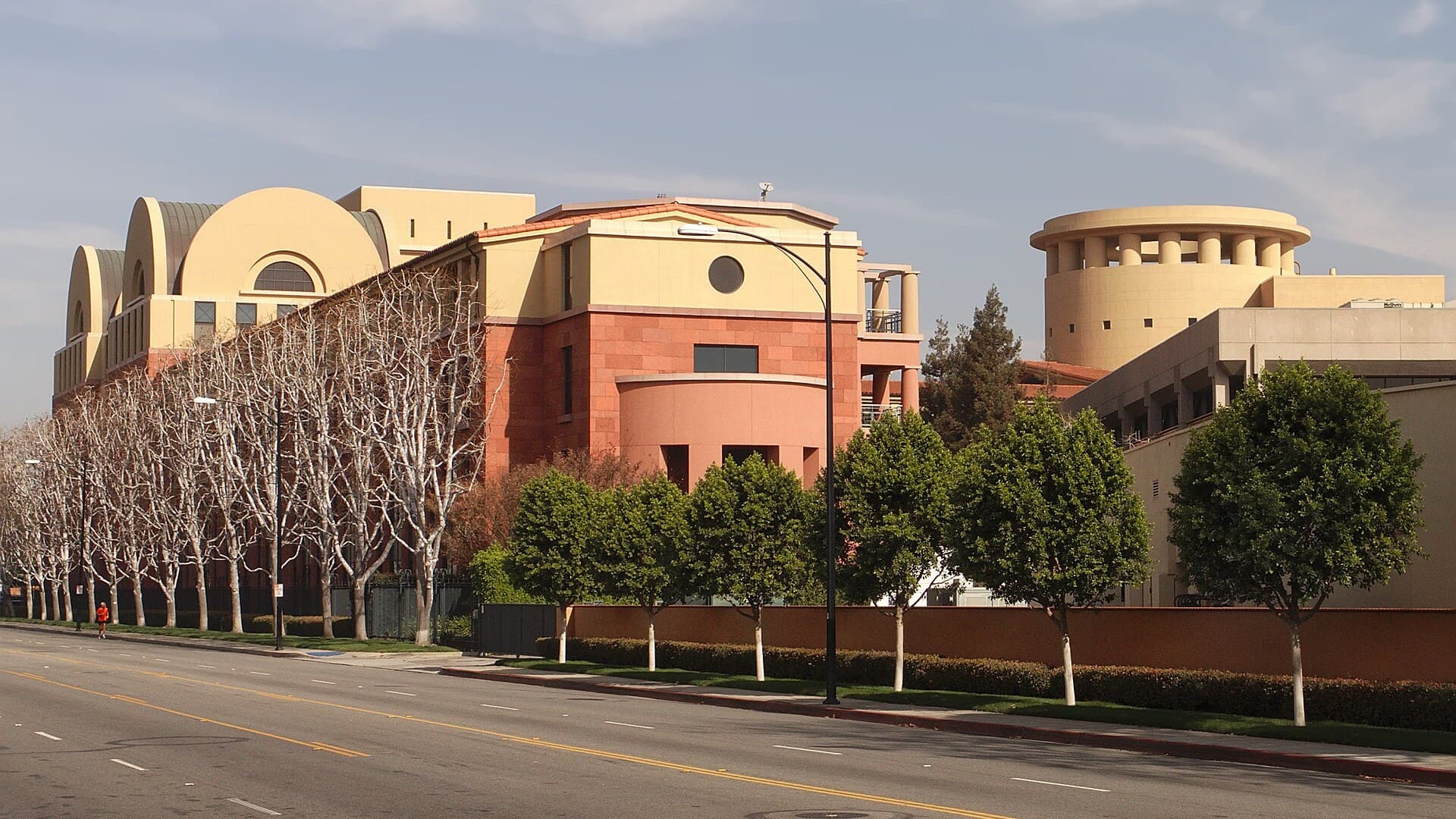 The Team Disney office building in Burbank, California, with its tan and terracotta facade and a circular tower, behind a row of trees along an empty street.