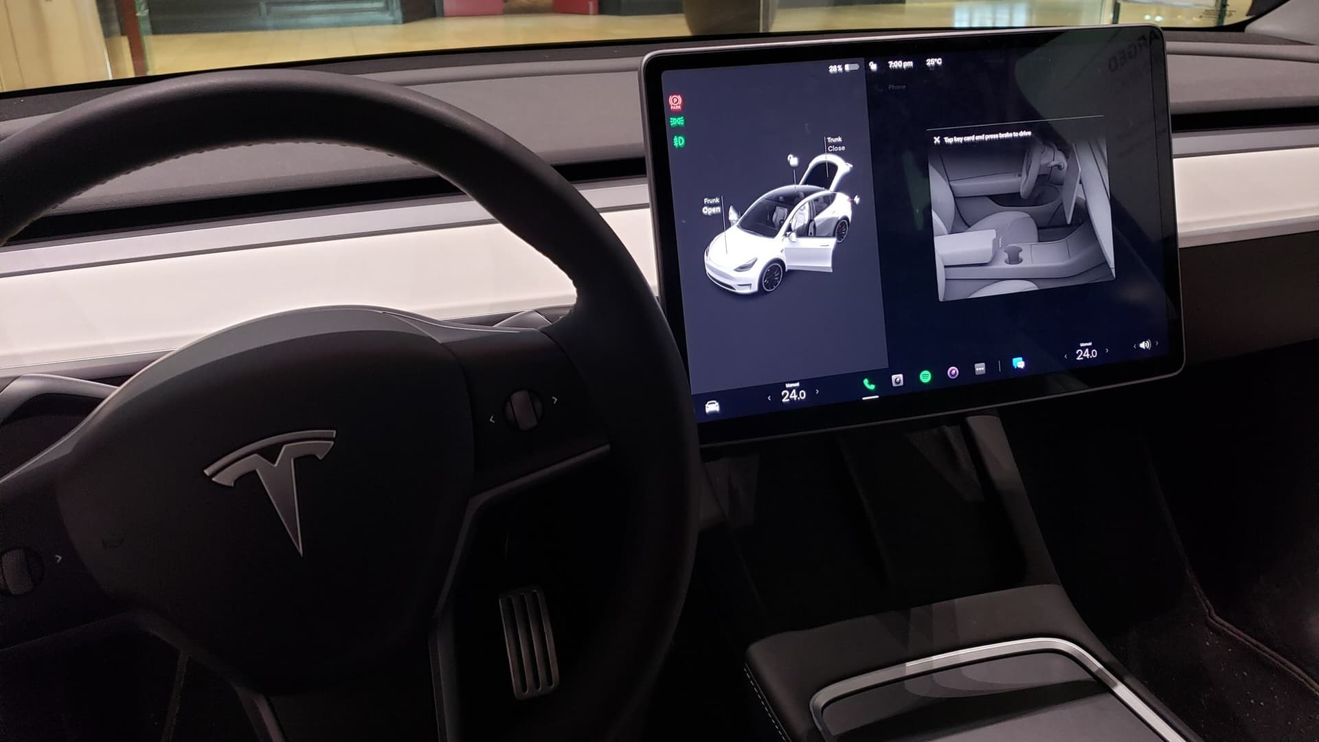 Inside a Tesla Model 3: a black steering wheel with the Tesla logo and a large central touchscreen showing a 3D image of the car with its trunk and frunk open.