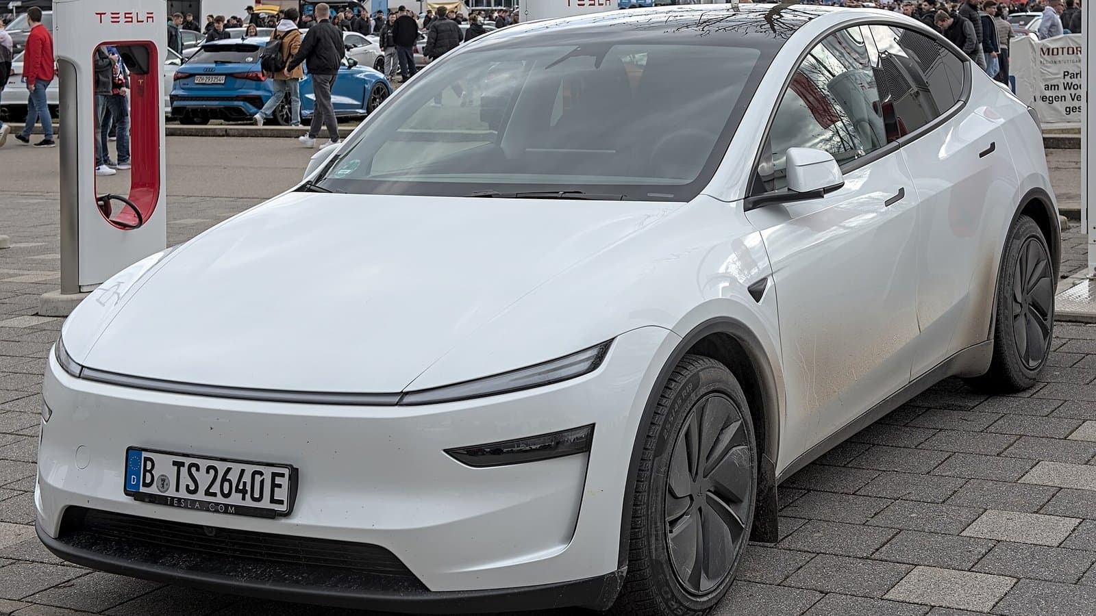A white redesigned Tesla Model Y electric SUV with a full-width light bar, parked near Tesla Superchargers.