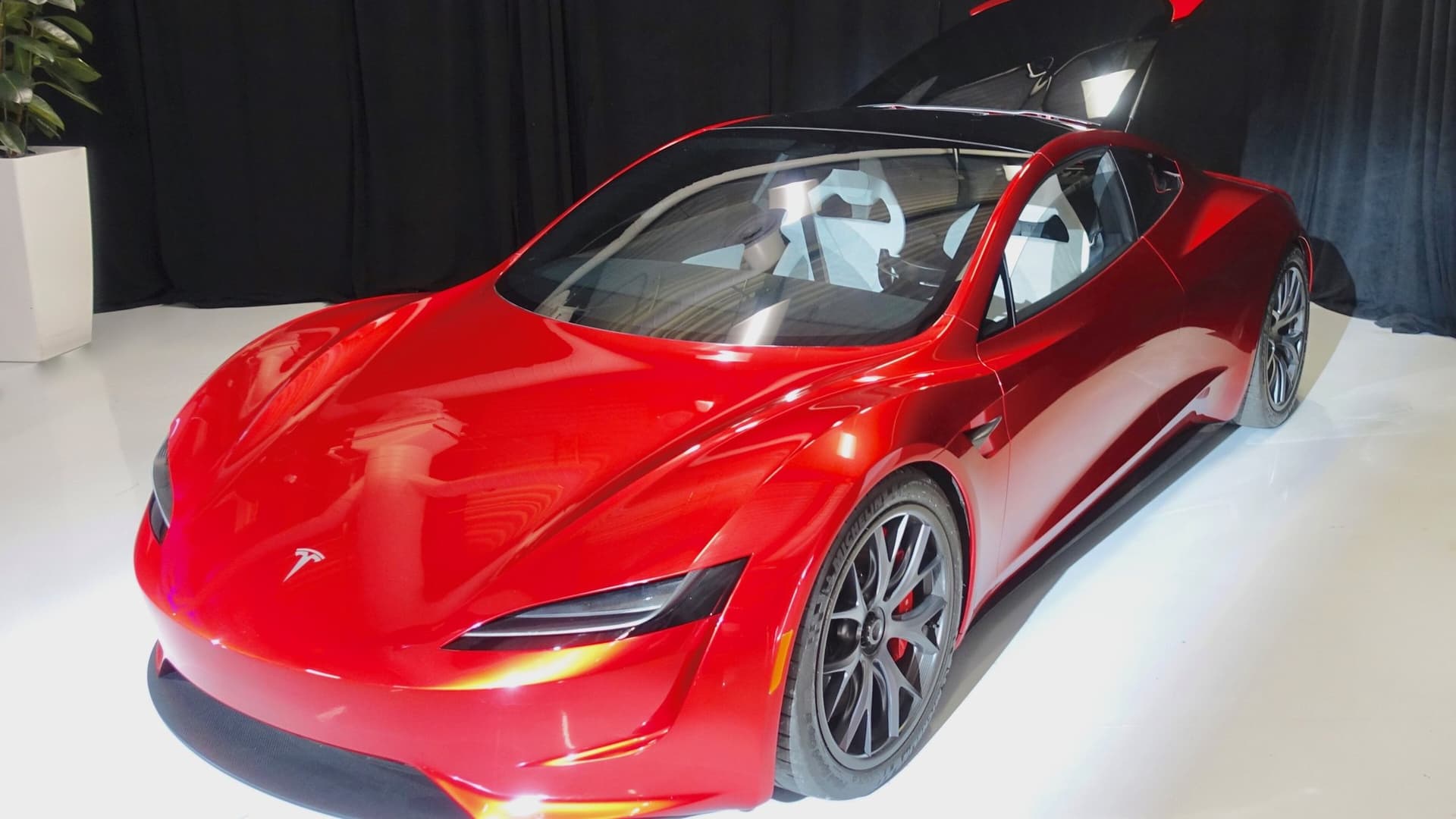 A glossy red Tesla Roadster prototype with a glass roof and silver wheels displayed indoors against a black curtain.