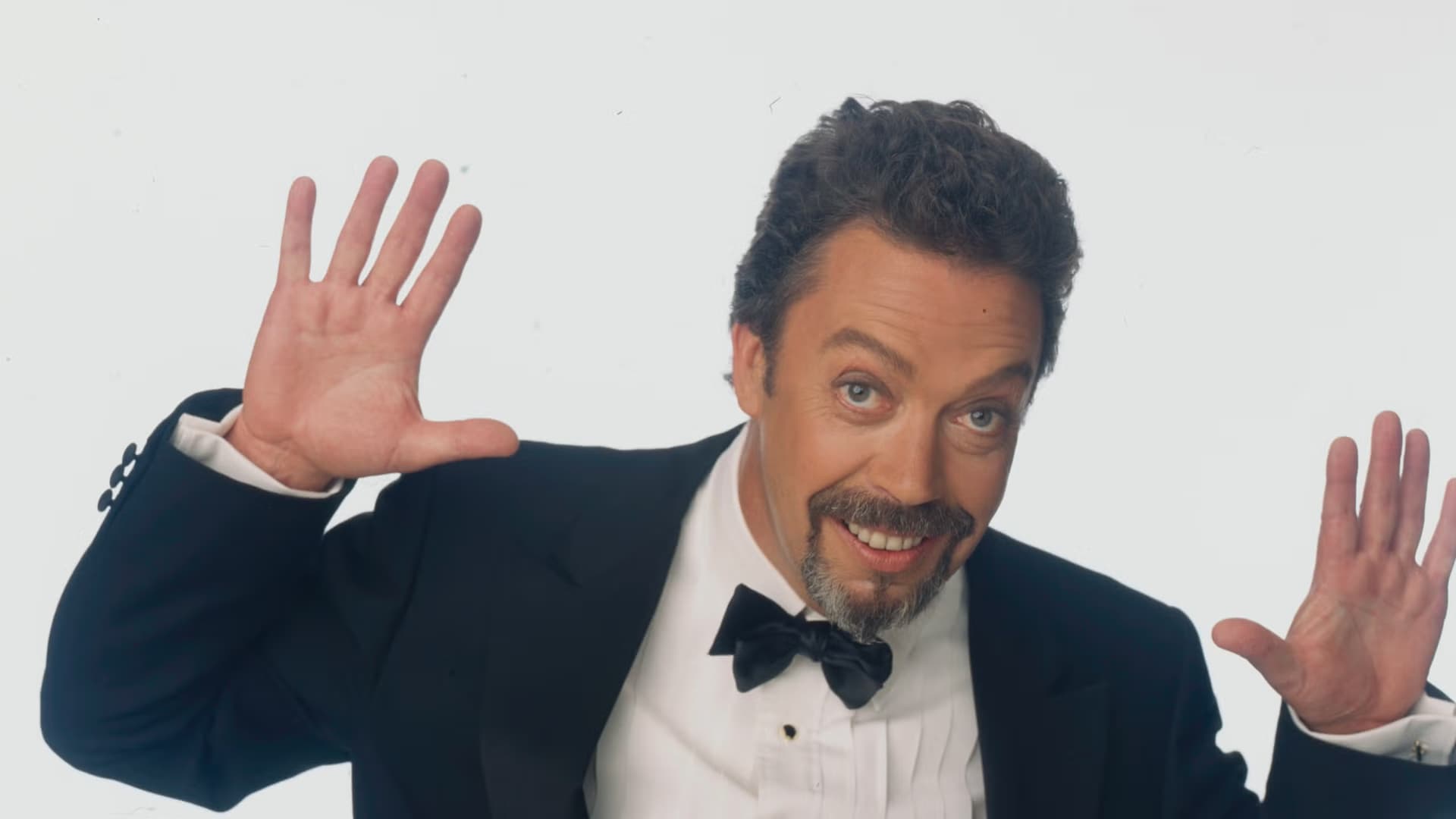 Tim Curry, in a dark suit, holding both hands up with a wry expression.